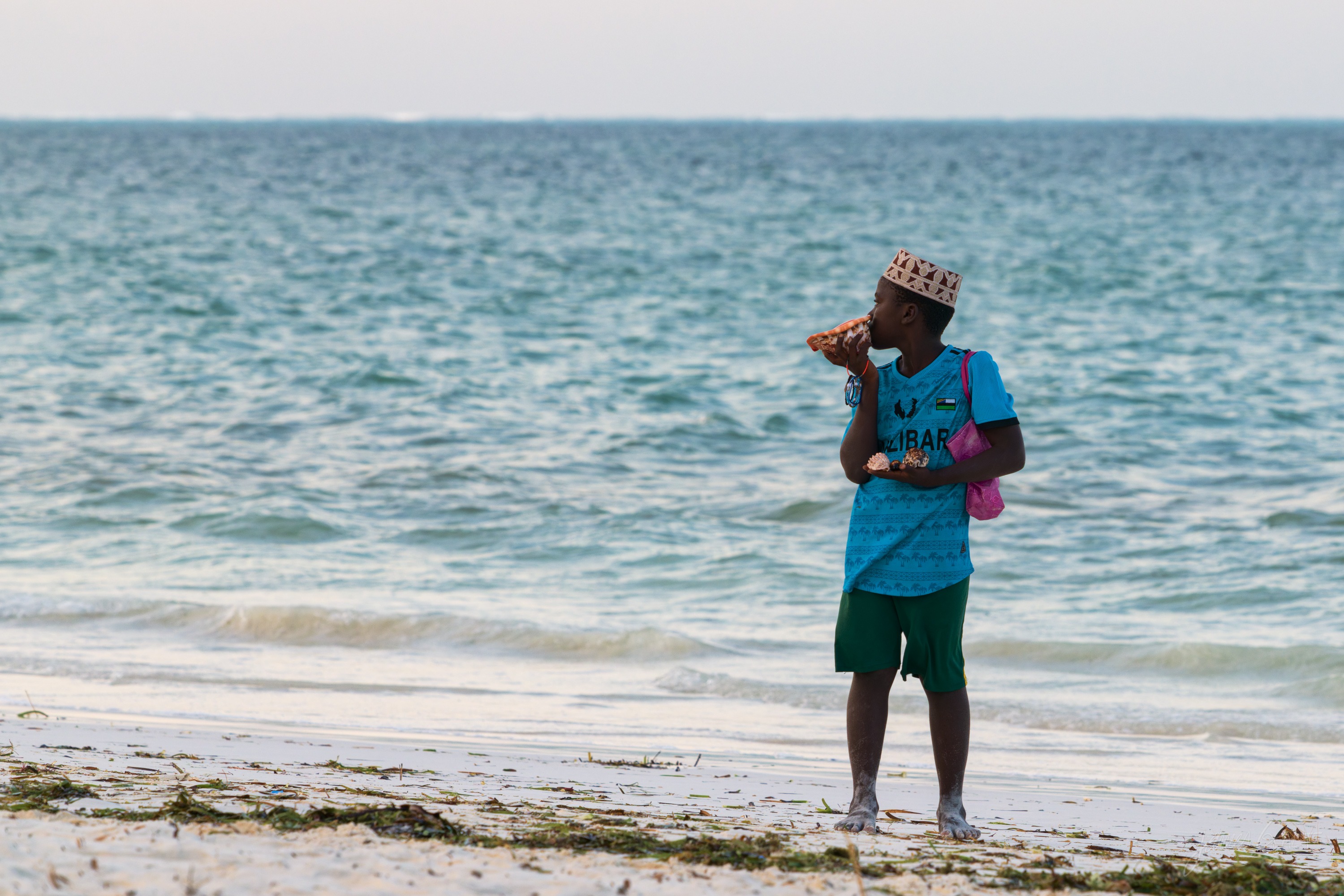 A man in a teal outfit walking along the wet sand carrying goods, the shallows and a hazy horizon behind him.
