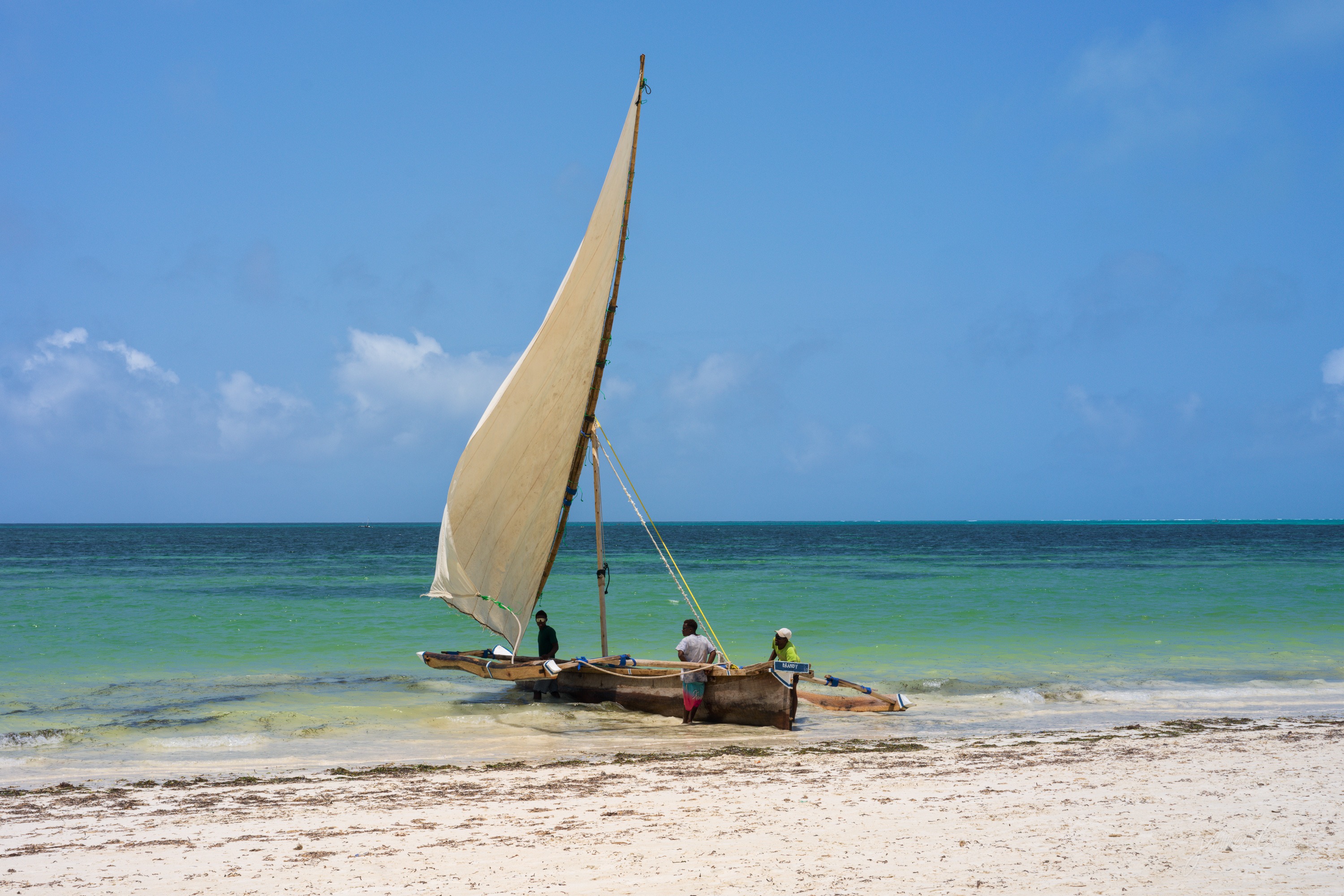 A wooden dhow drawn up on a white-sand beach with its triangular sail still raised, turquoise shallows and a pale sky behind it.