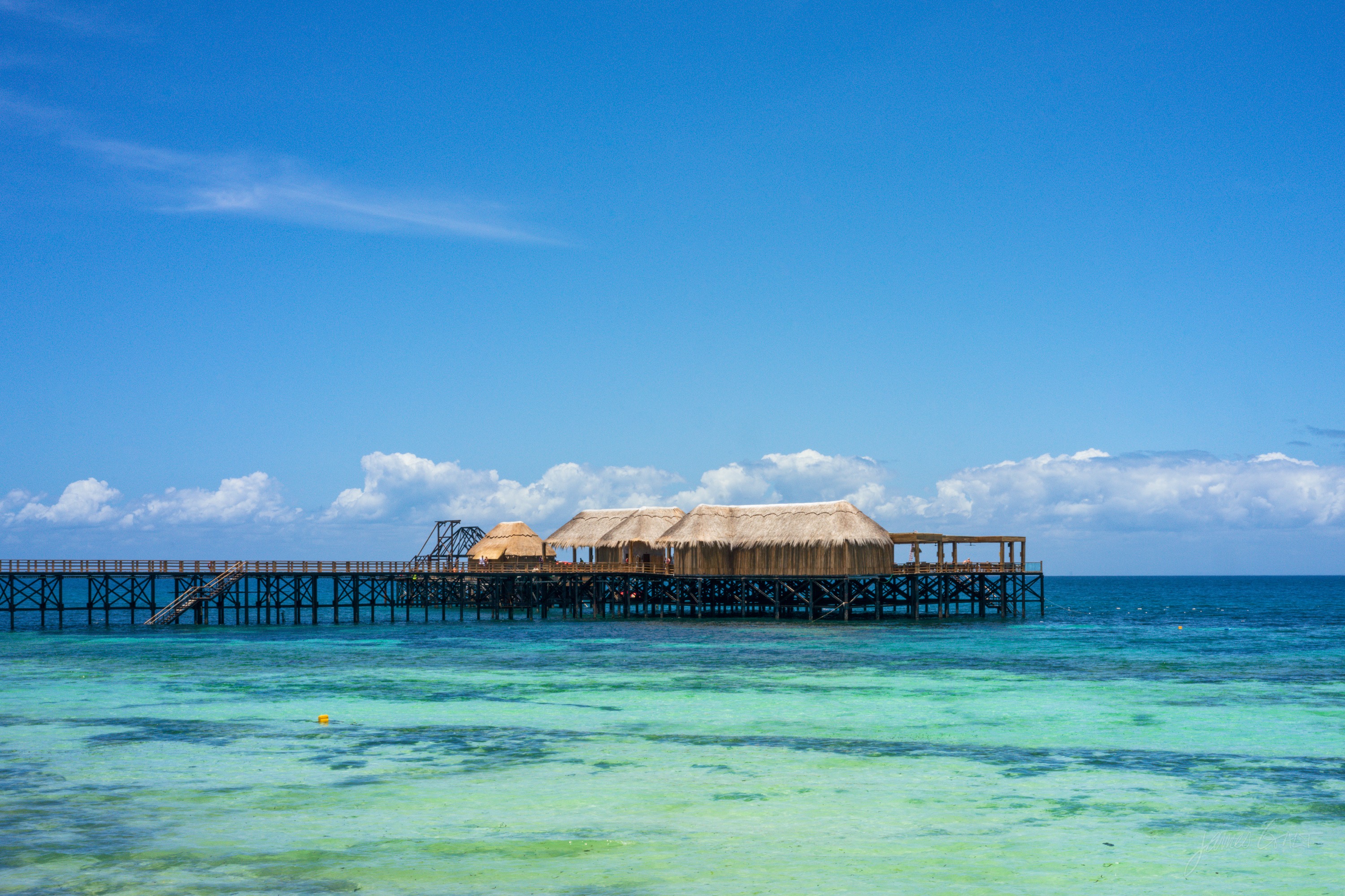 A small thatched bar built on stilts out over clear turquoise water, reached by a low walkway, a few figures on it under a bright sky.