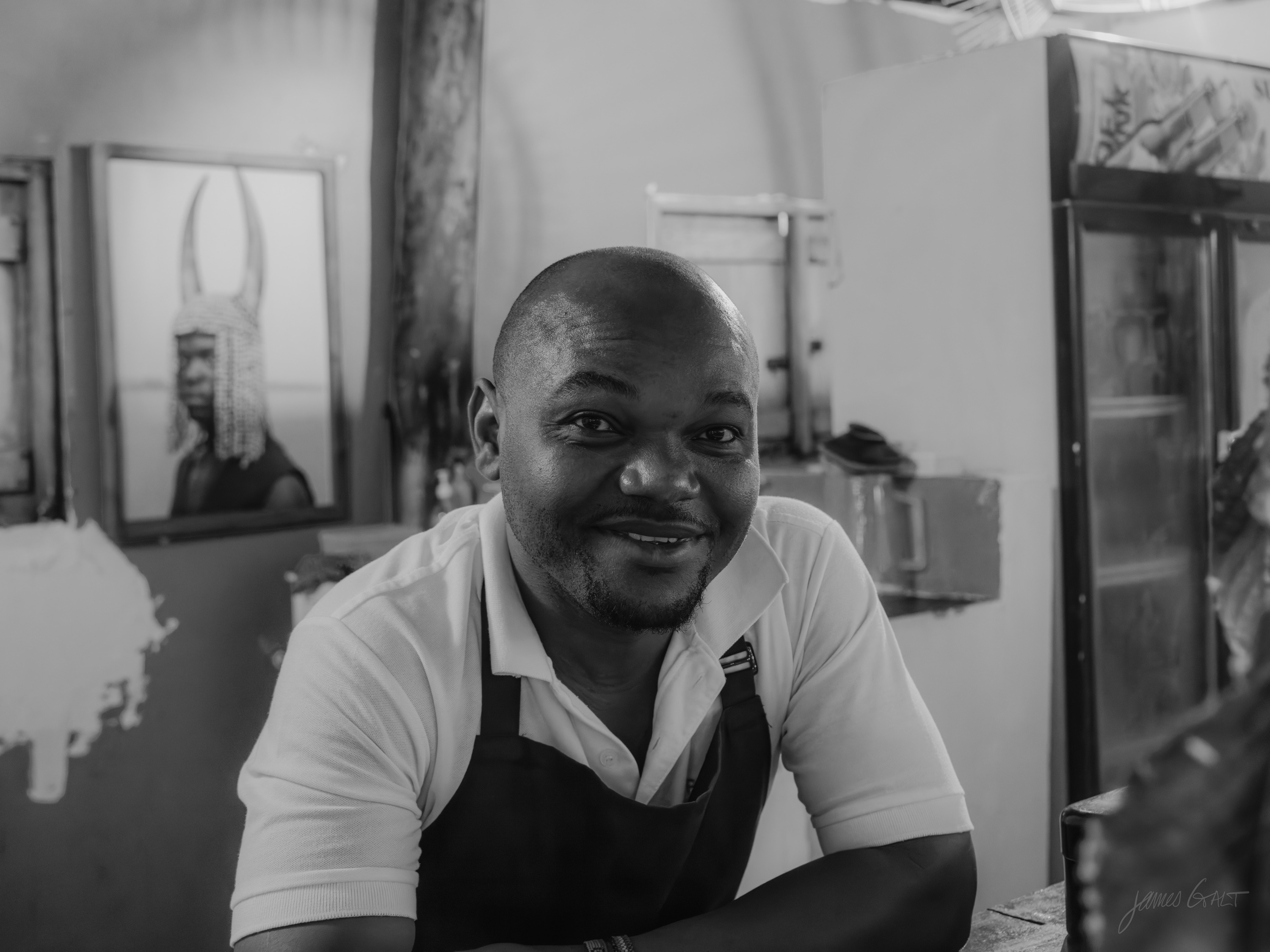 Black and white portrait of a smiling man in a white shirt and apron standing in a kitchen or bar, a framed picture on the wall behind him.