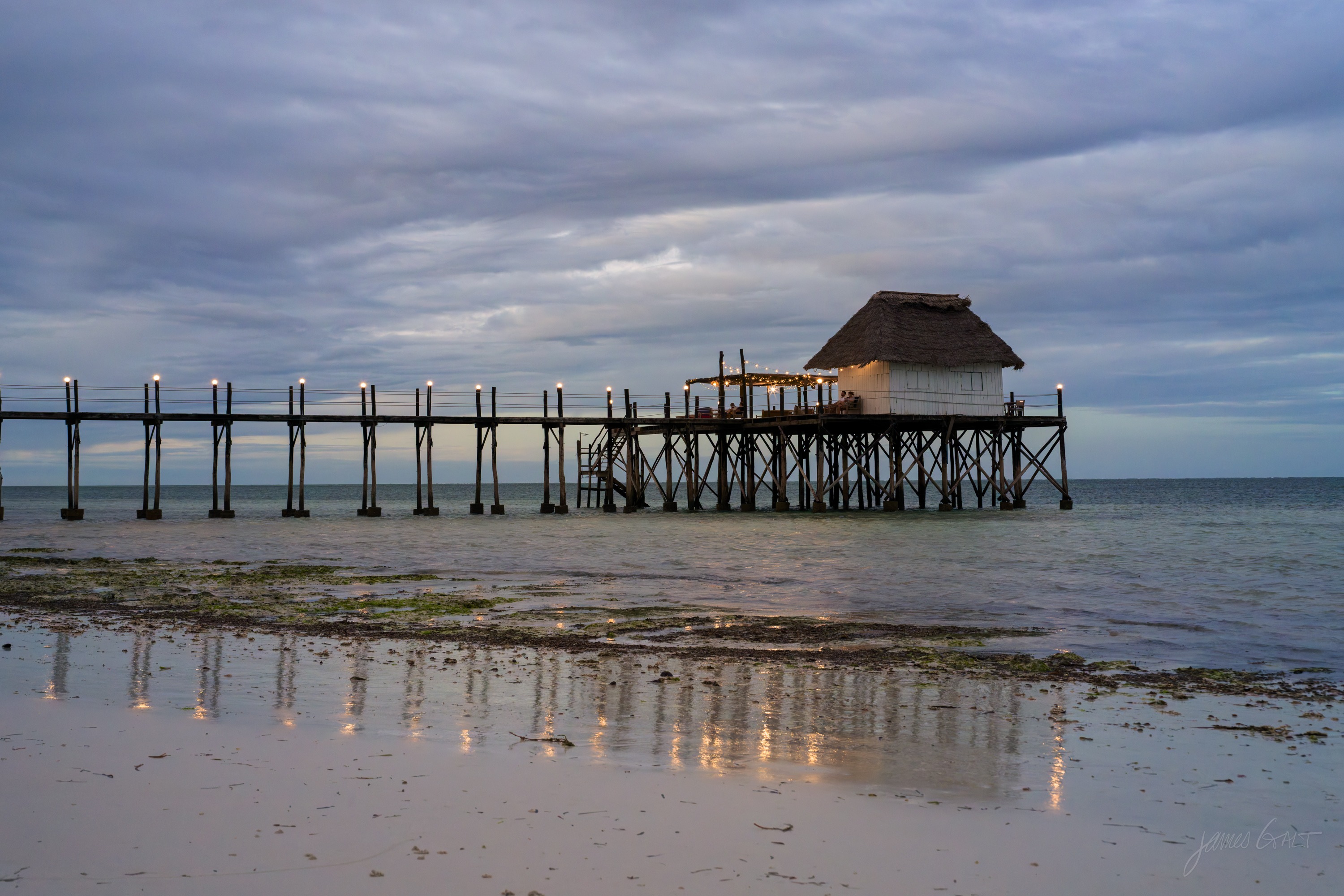 A long wooden pier in silhouette against a dusky sky, its legs reflected in the wet sand, the flat sea behind.