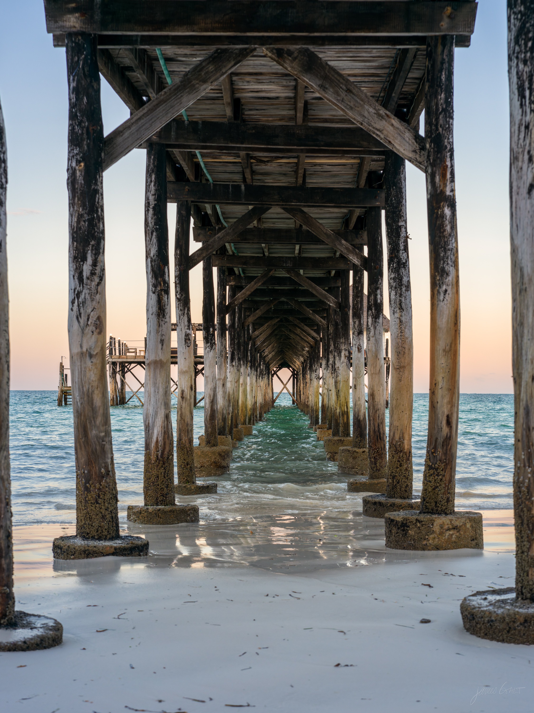 A view along the underside of a pier, rows of wooden pilings receding toward a bright opening of sea and sky at the far end.
