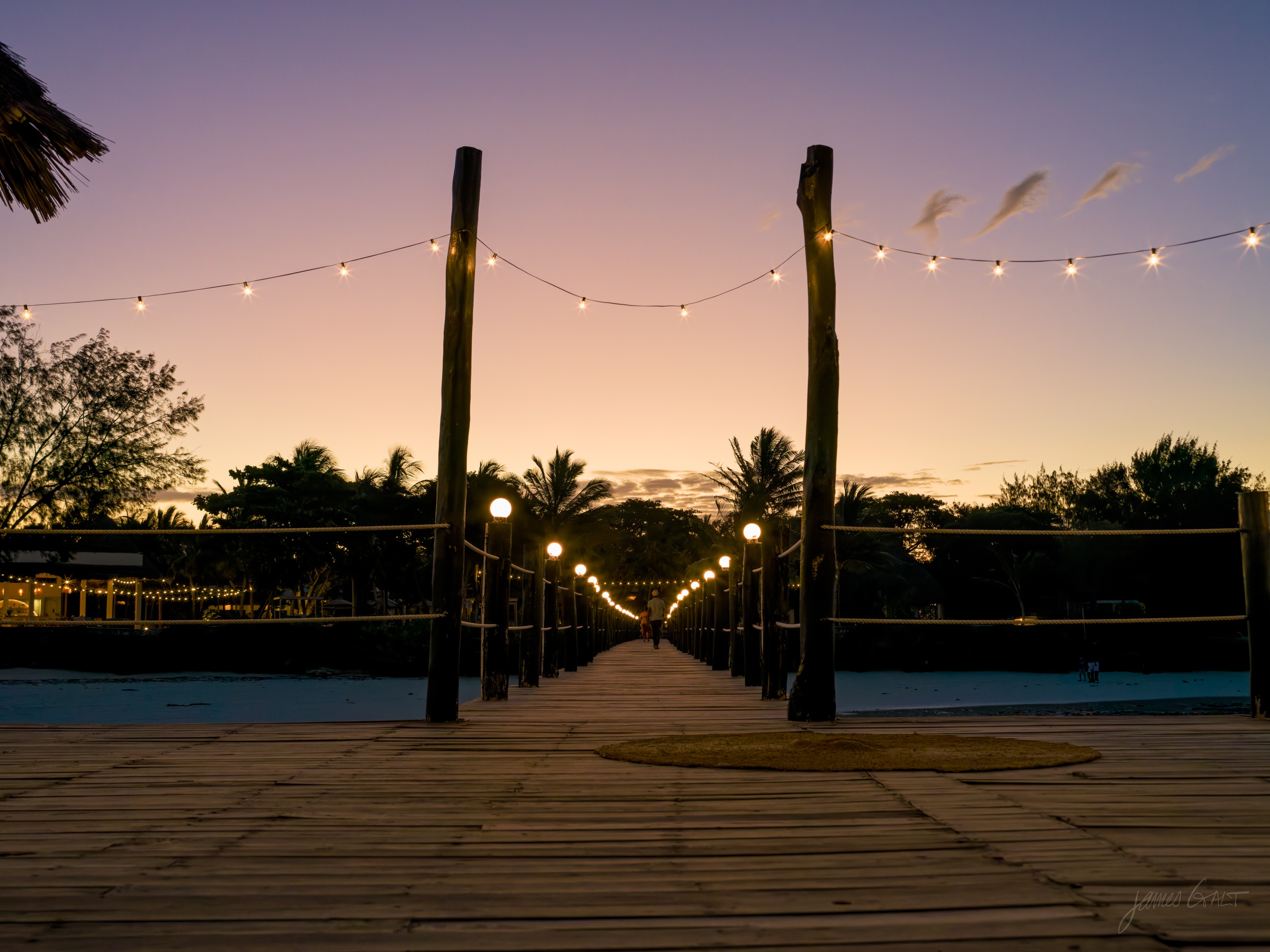 A pier or boardwalk strung with small warm lights at dusk, receding toward the water, the sky deep blue above.
