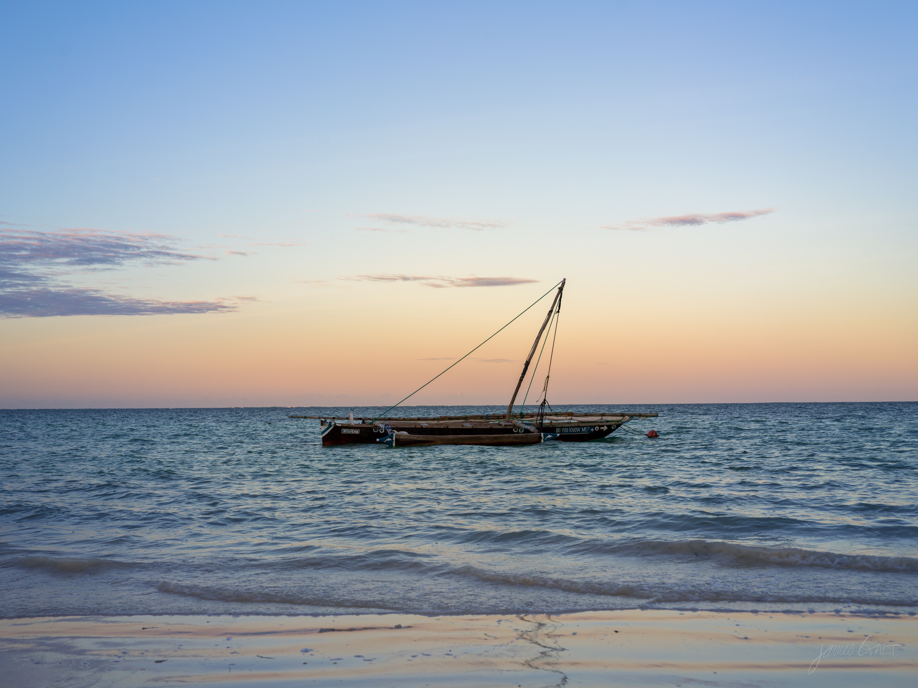 A dhow anchored offshore in colour at sunset, its bare mast against a band of warm sky, calm water in the foreground.