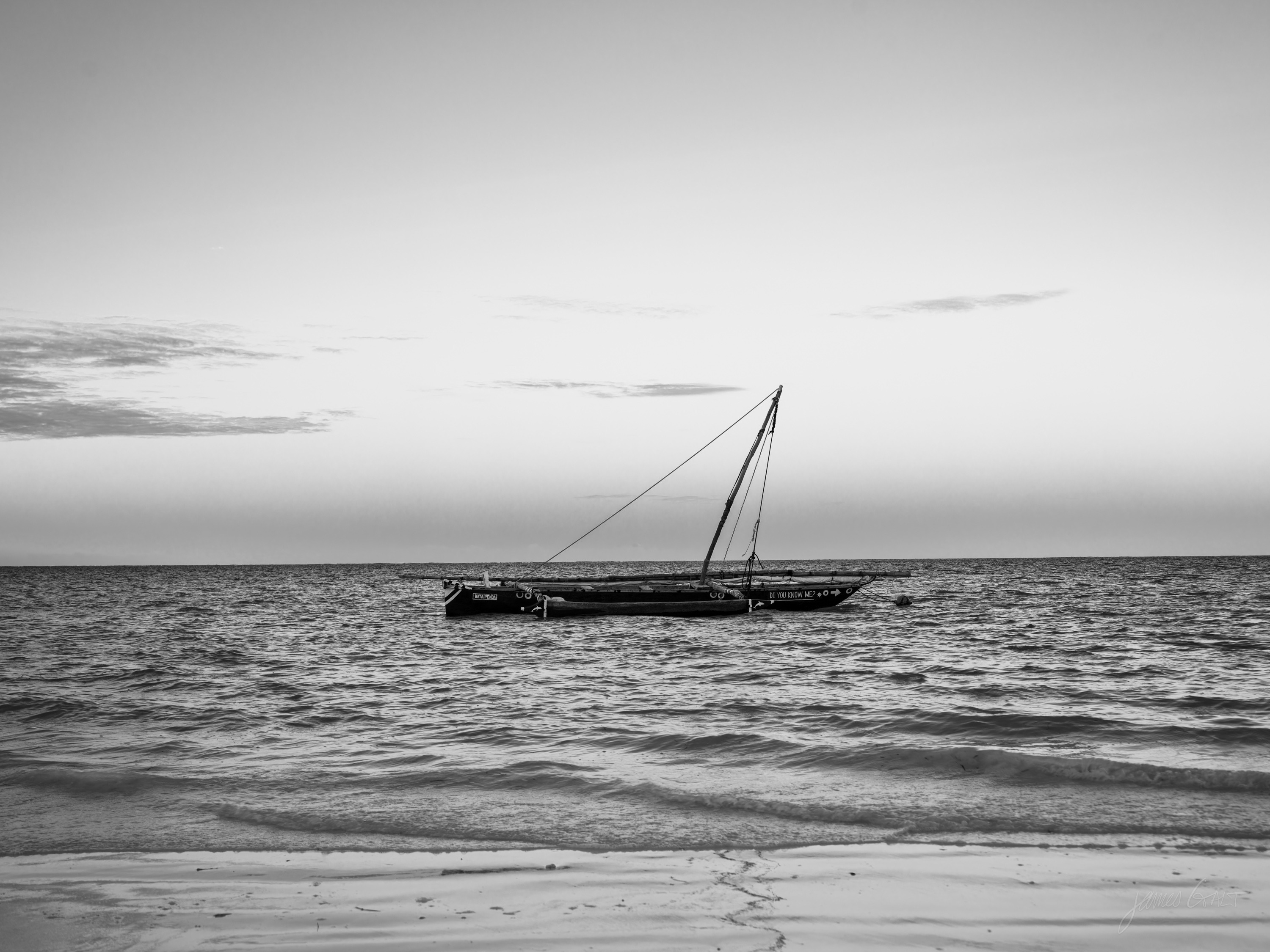 The same dhow in black and white, a dark silhouette on flat water beneath a bright, textured sky.