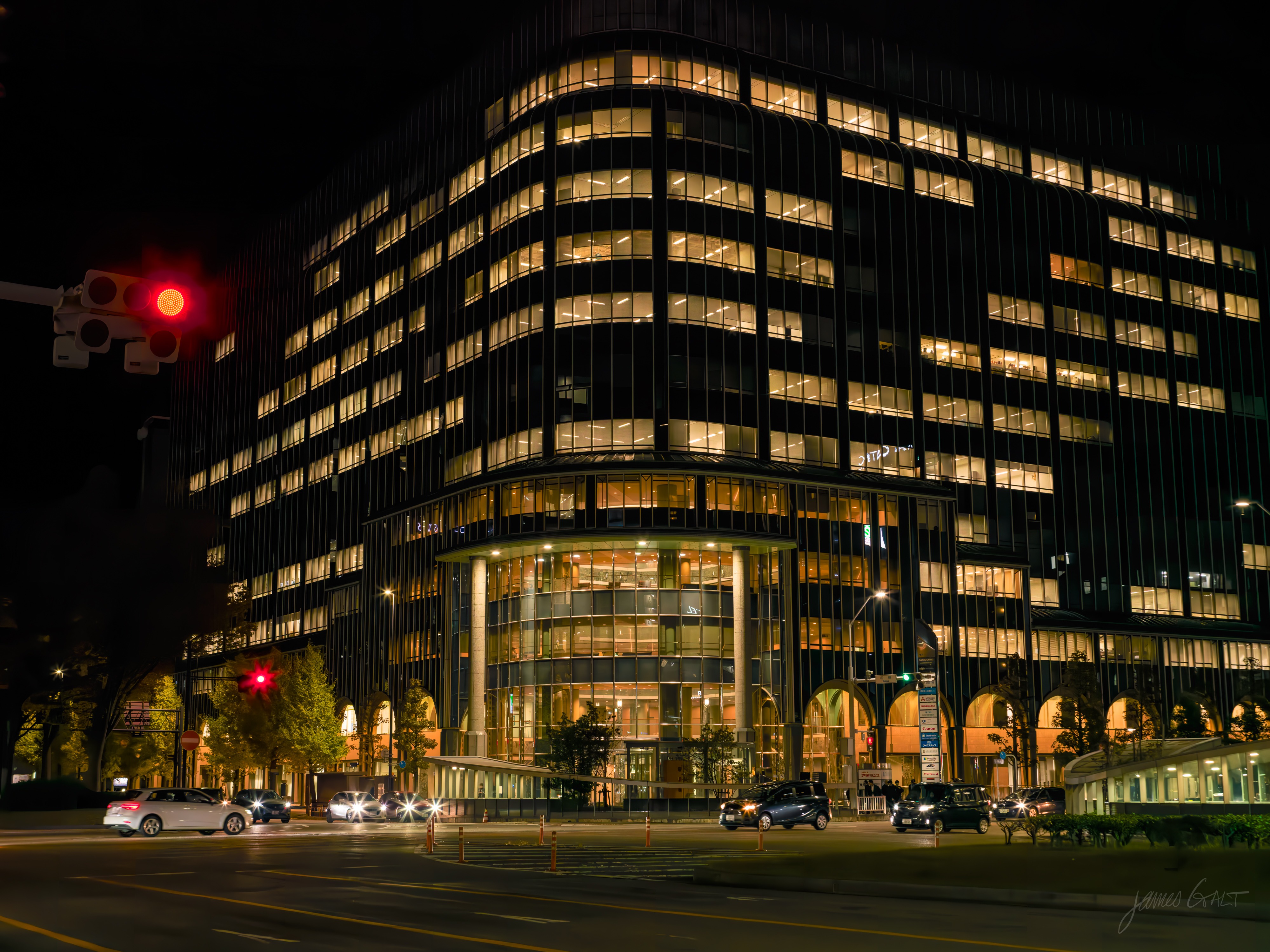 A large modern office building at night with a curved glass corner and rows of lit windows, a warm-lit lobby and arched colonnade at its base. A red traffic light and passing cars in the foreground at a wide intersection.