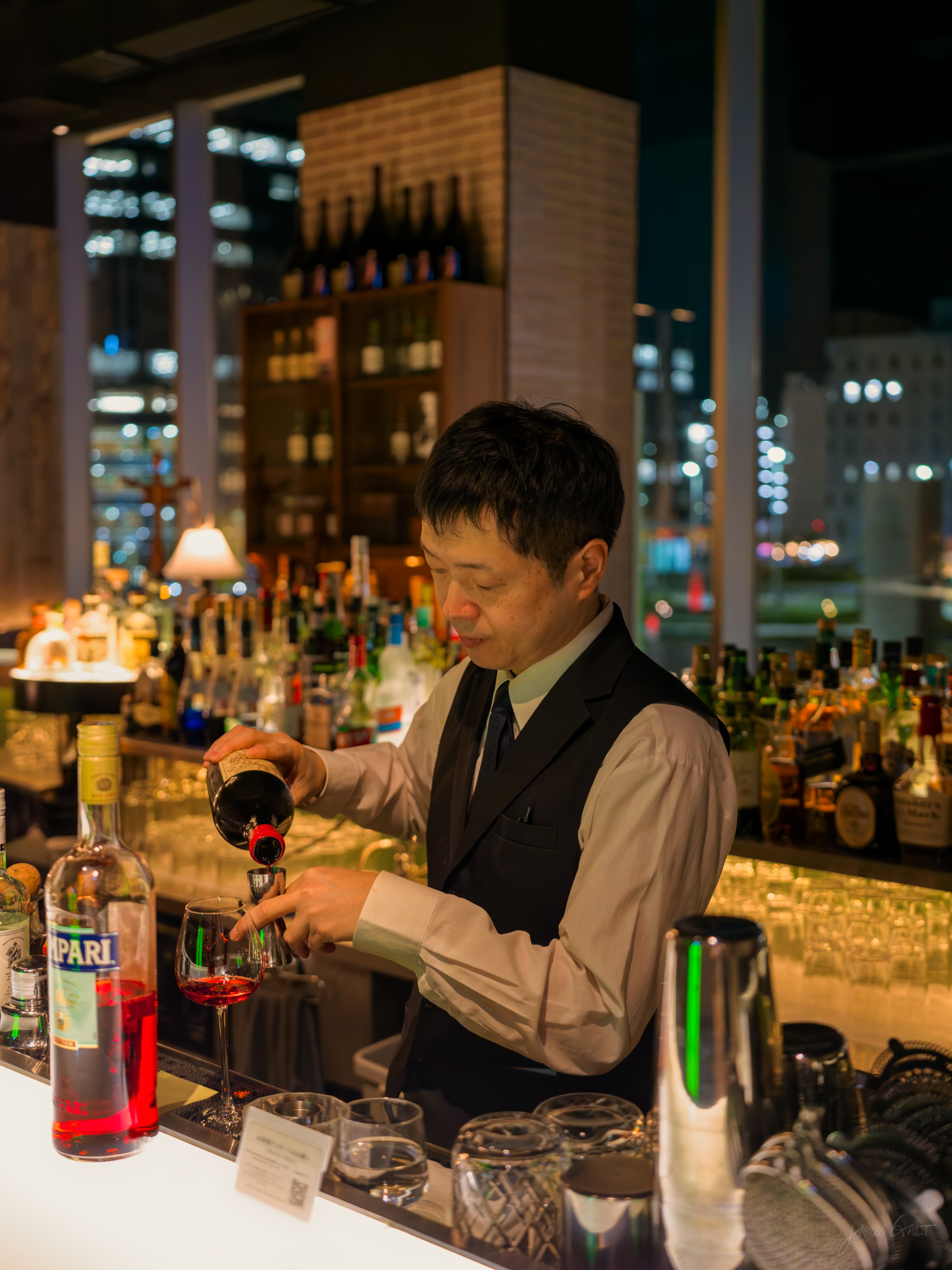 A bartender in a waistcoat and tie pours a dark red liqueur into a glass at a hotel bar, a Campari bottle in front of him and shelves of spirits around, the lights of the city visible through a window behind.