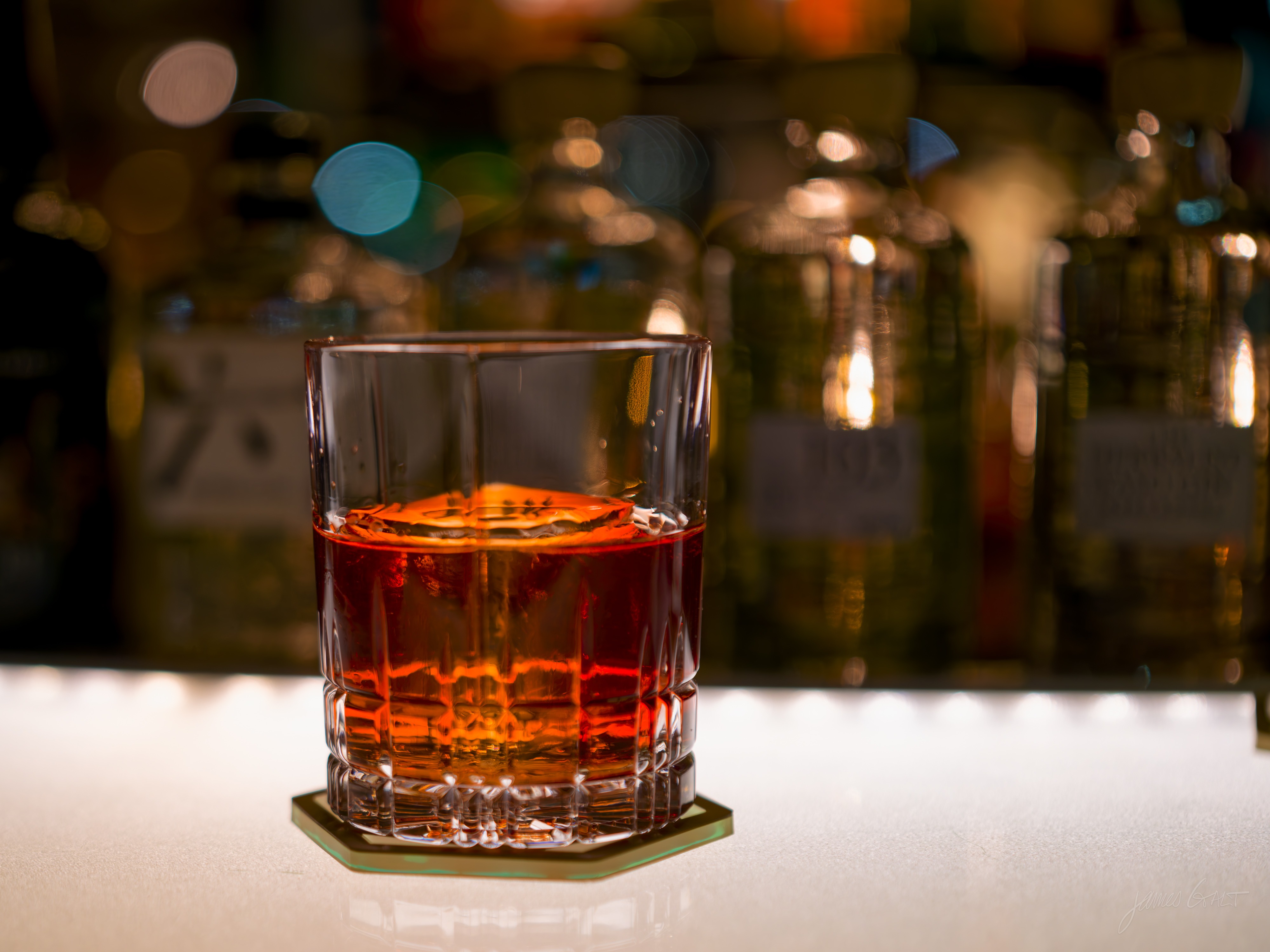 A negroni, deep red over ice in a faceted tumbler on a coaster, lit warm on a pale bar top with rows of backlit bottles softly out of focus behind it.