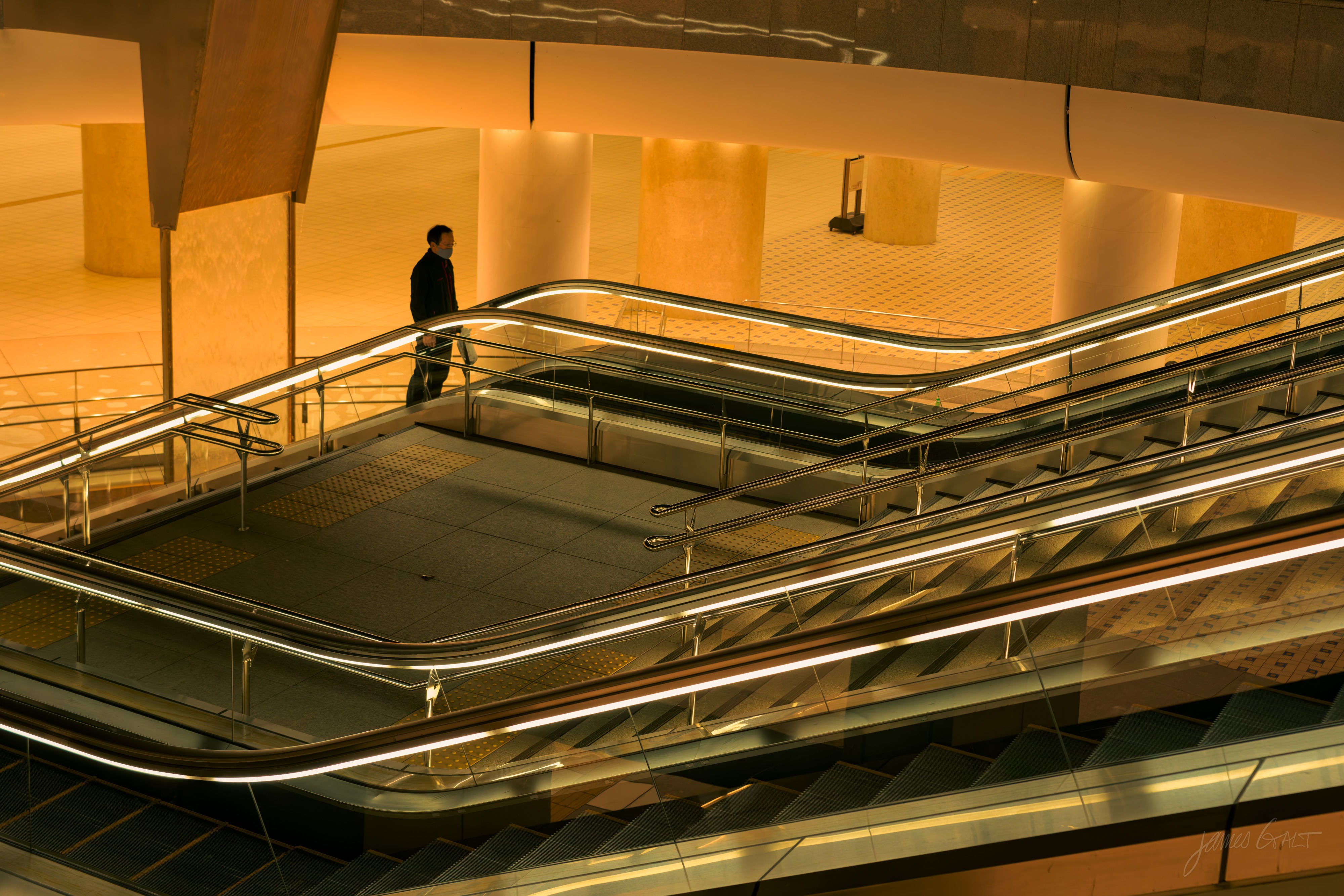 A modern interior atrium lit warm gold, with criss-crossing escalators and curved metal handrails, a single person walking along a walkway at the top.