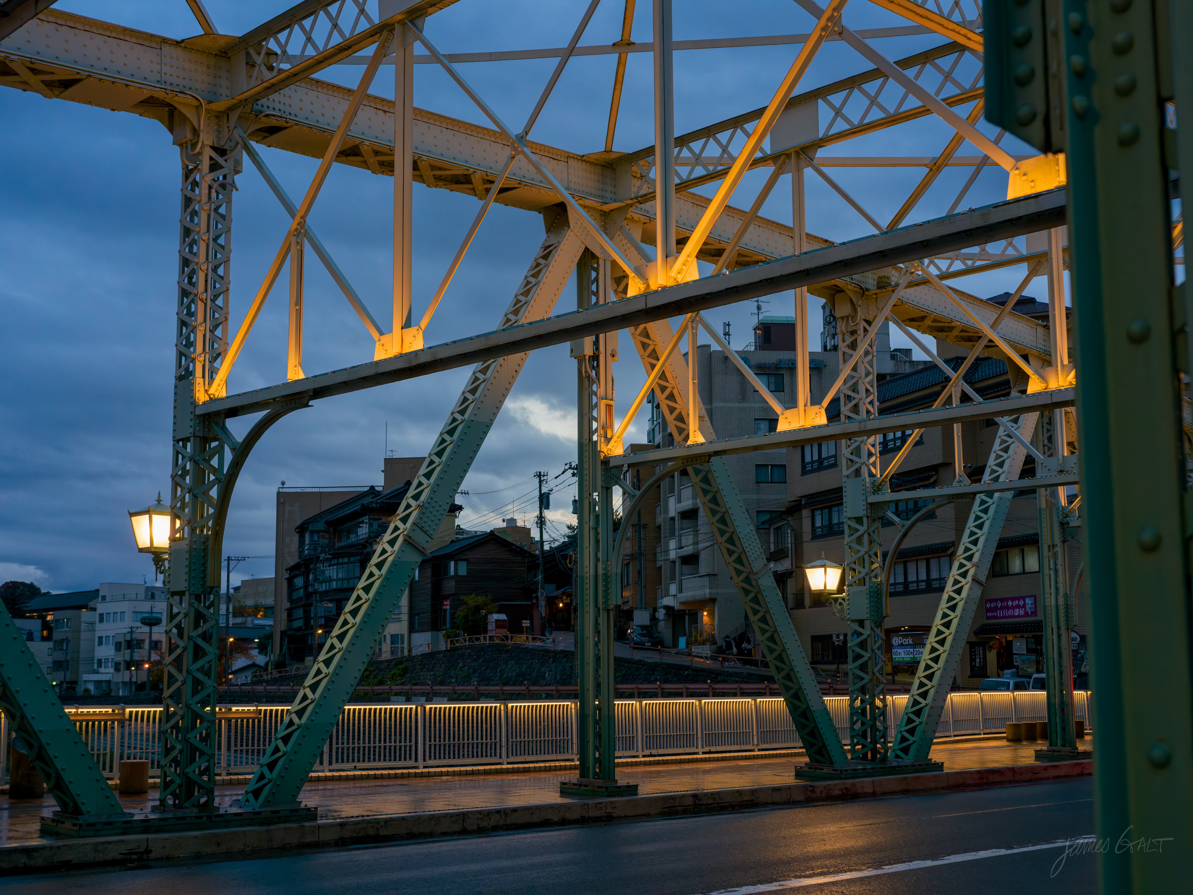 A green-painted steel truss bridge at dusk, its girders lit warm amber from below against a deep blue sky. Retro lamp posts with glowing white globes line the deck, low town buildings sit beyond, and the road is wet underfoot.