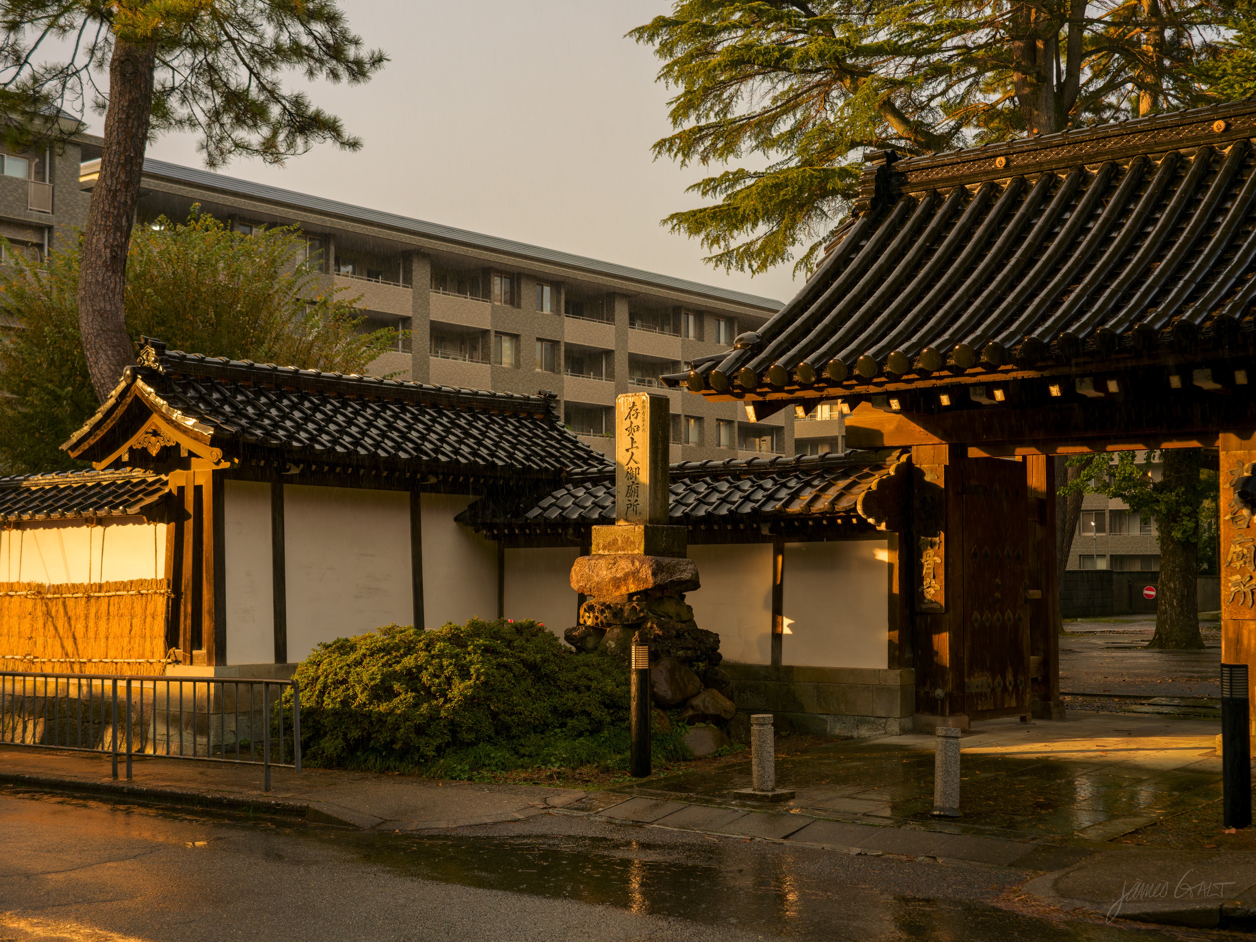 A traditional gate and white-walled building with dark tiled roofs in warm evening light, a tall inscribed stone marker between them and a modern apartment block behind. The road in front is wet from rain.