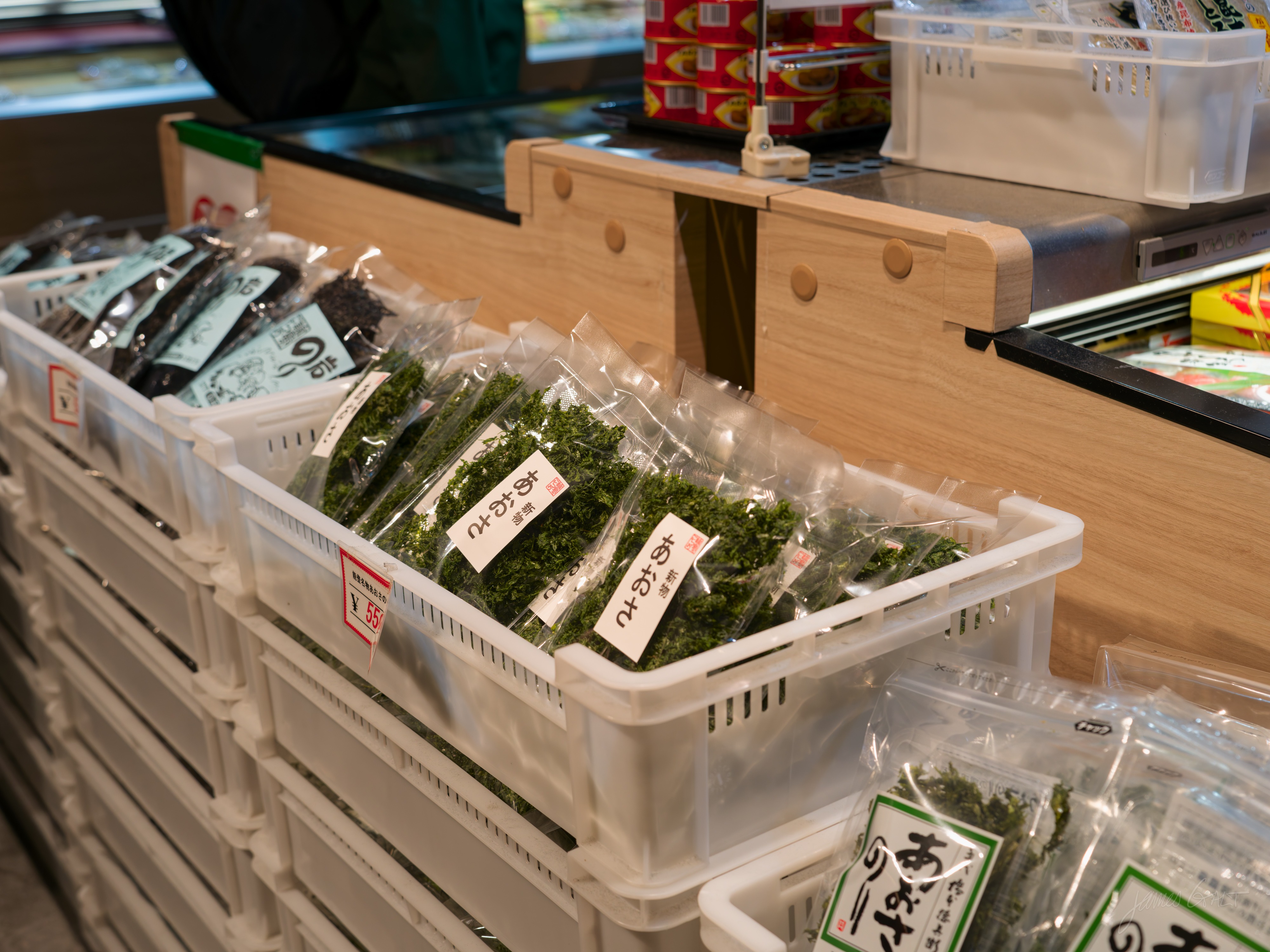 Close-up of a market stall's crates: clear plastic packs of green seaweed and dark dried nori with small price labels, stacked in white baskets on a wooden counter.