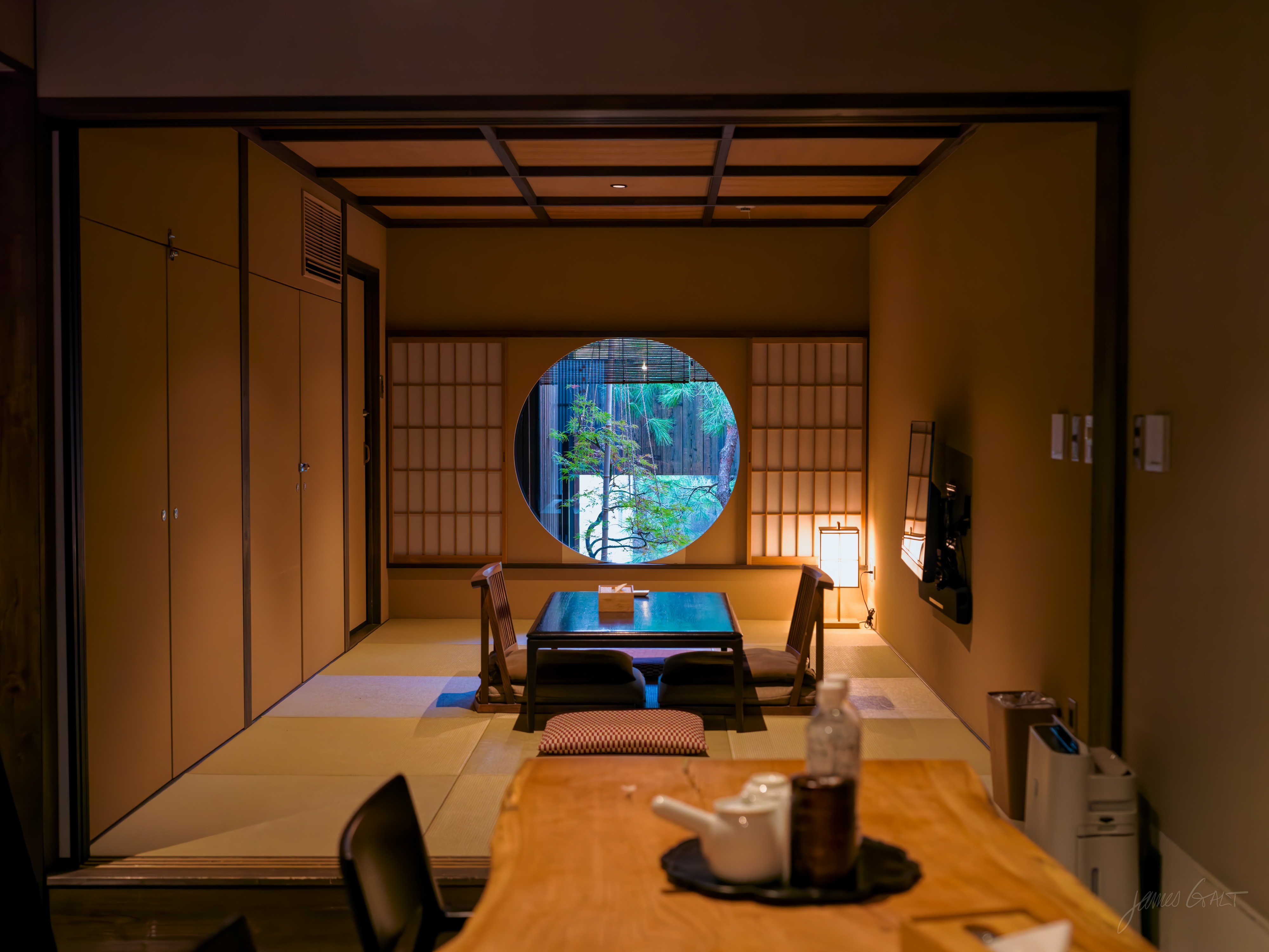 The interior of a traditional Japanese inn room at dusk: tatami floor, shoji screens, a low table with legless chairs and a glowing paper lamp. In the back wall a large round window frames a single pine branch against a small garden going dark outside. A wooden table with a teapot sits in the warm-lit foreground.