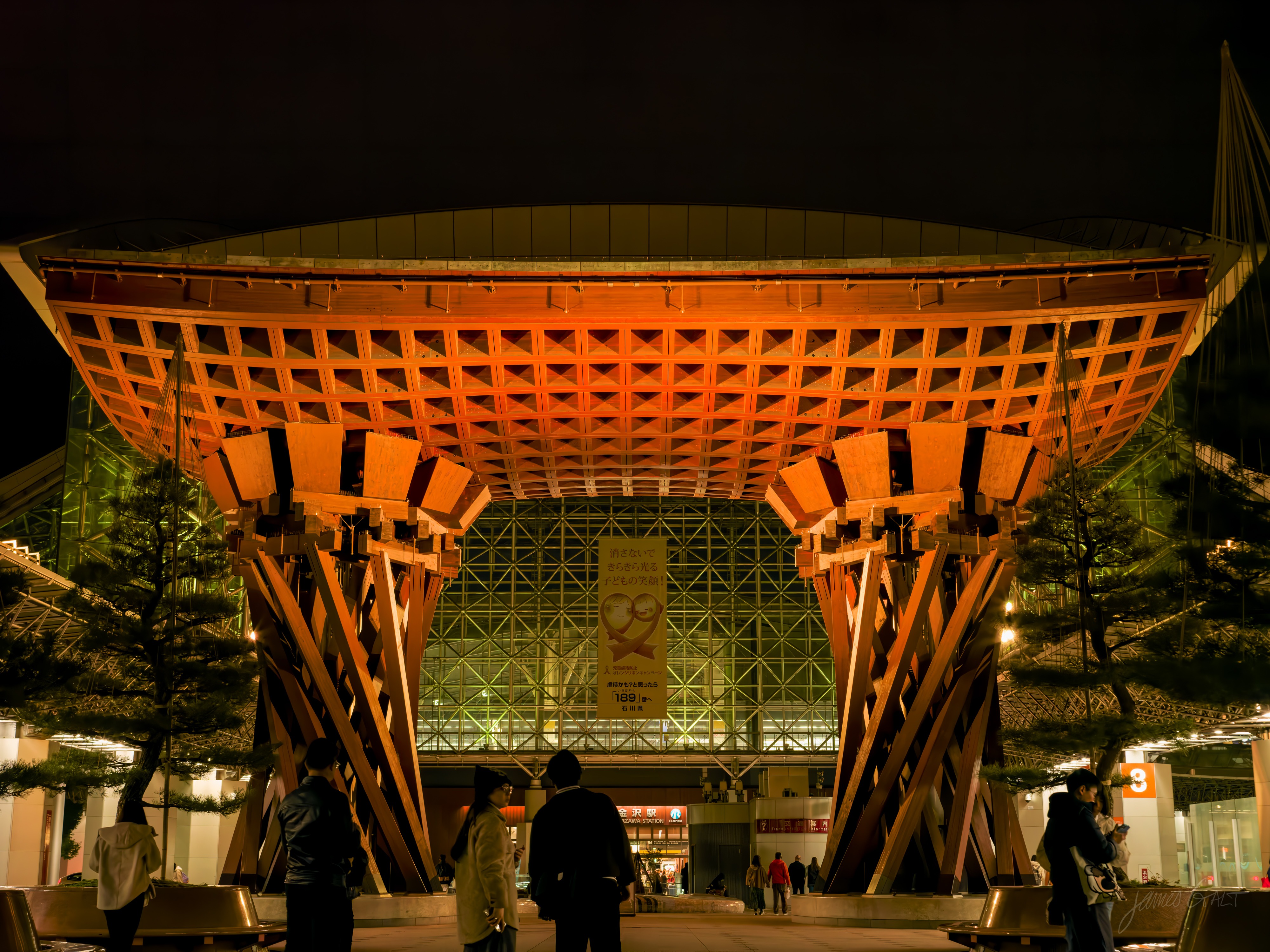 The Tsuzumi-mon gate at Kanazawa Station at night, a massive wooden gate of crossed timber lattice lit warm orange, standing in front of the station's large glass-and-steel dome. Several people stand silhouetted beneath it.