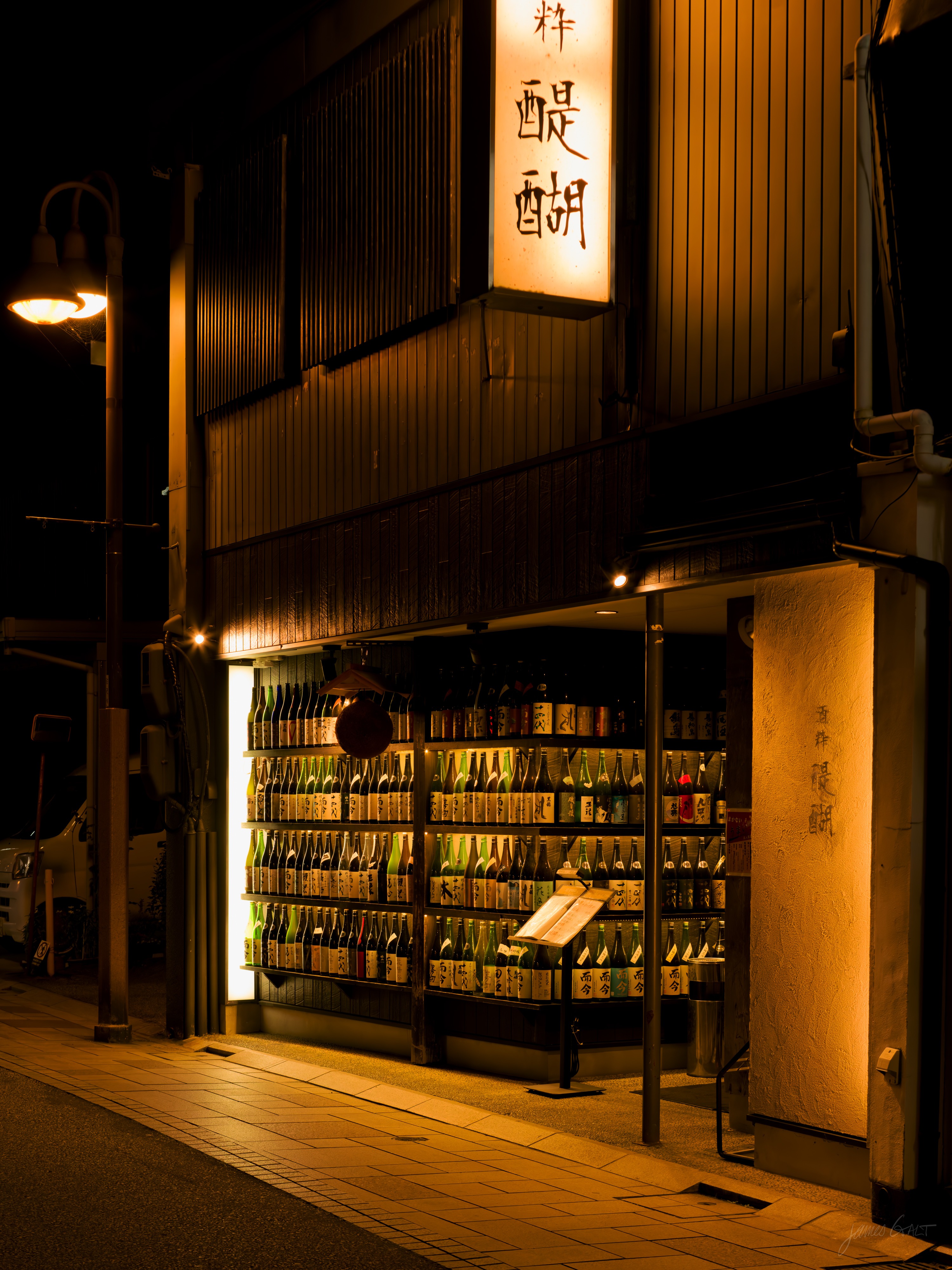 A traditional wooden sake shop at night, its open frontage glowing with backlit shelves packed full of sake bottles. A vertical illuminated sign with Japanese characters hangs above, a street lamp and quiet road to the left.