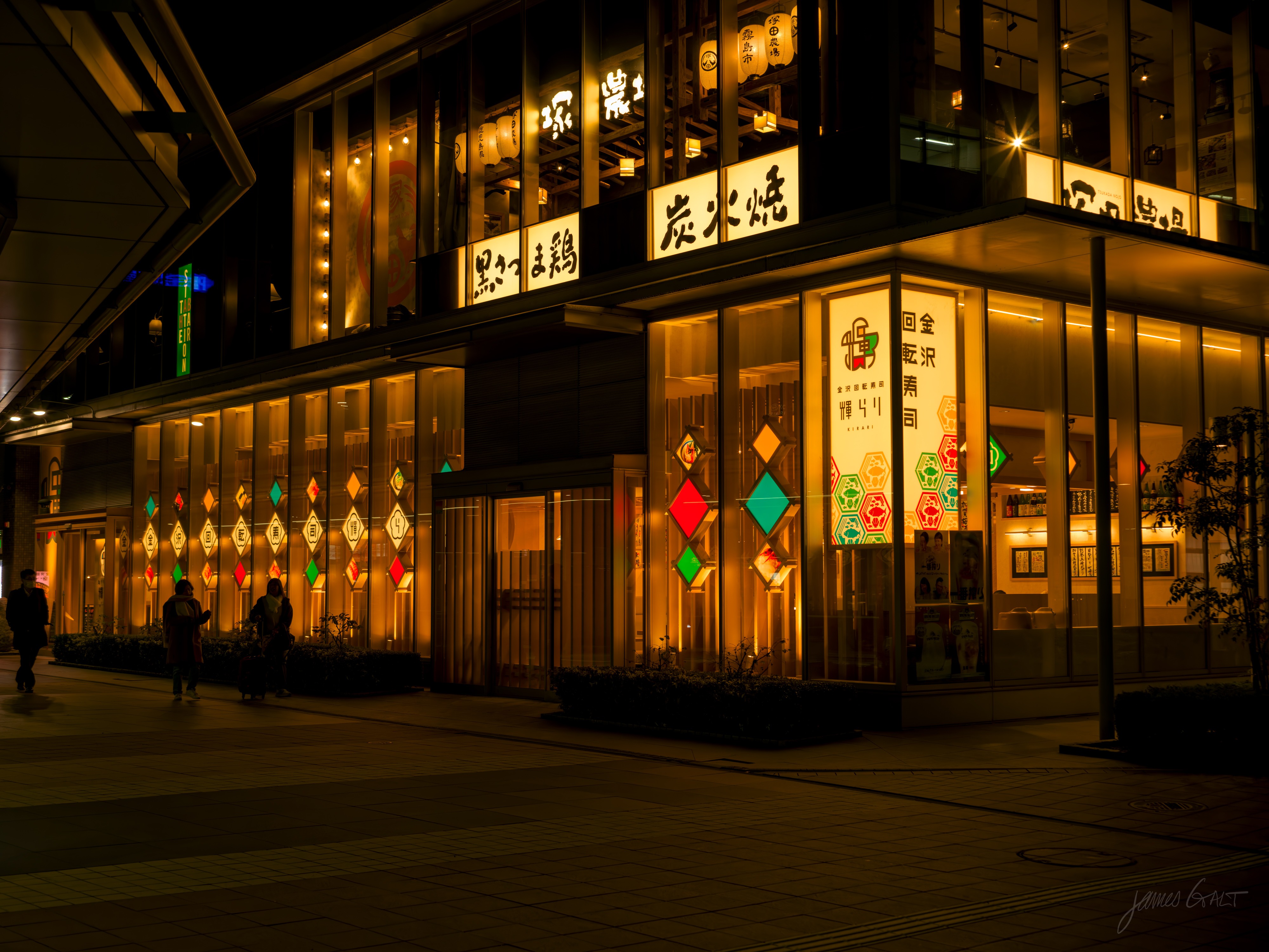 A restaurant building at night with large windows set with colourful diamond-patterned glass in red, green and yellow, warm light glowing from inside. Japanese signs for charcoal grill run along the upper facade, a few people on the plaza in front.