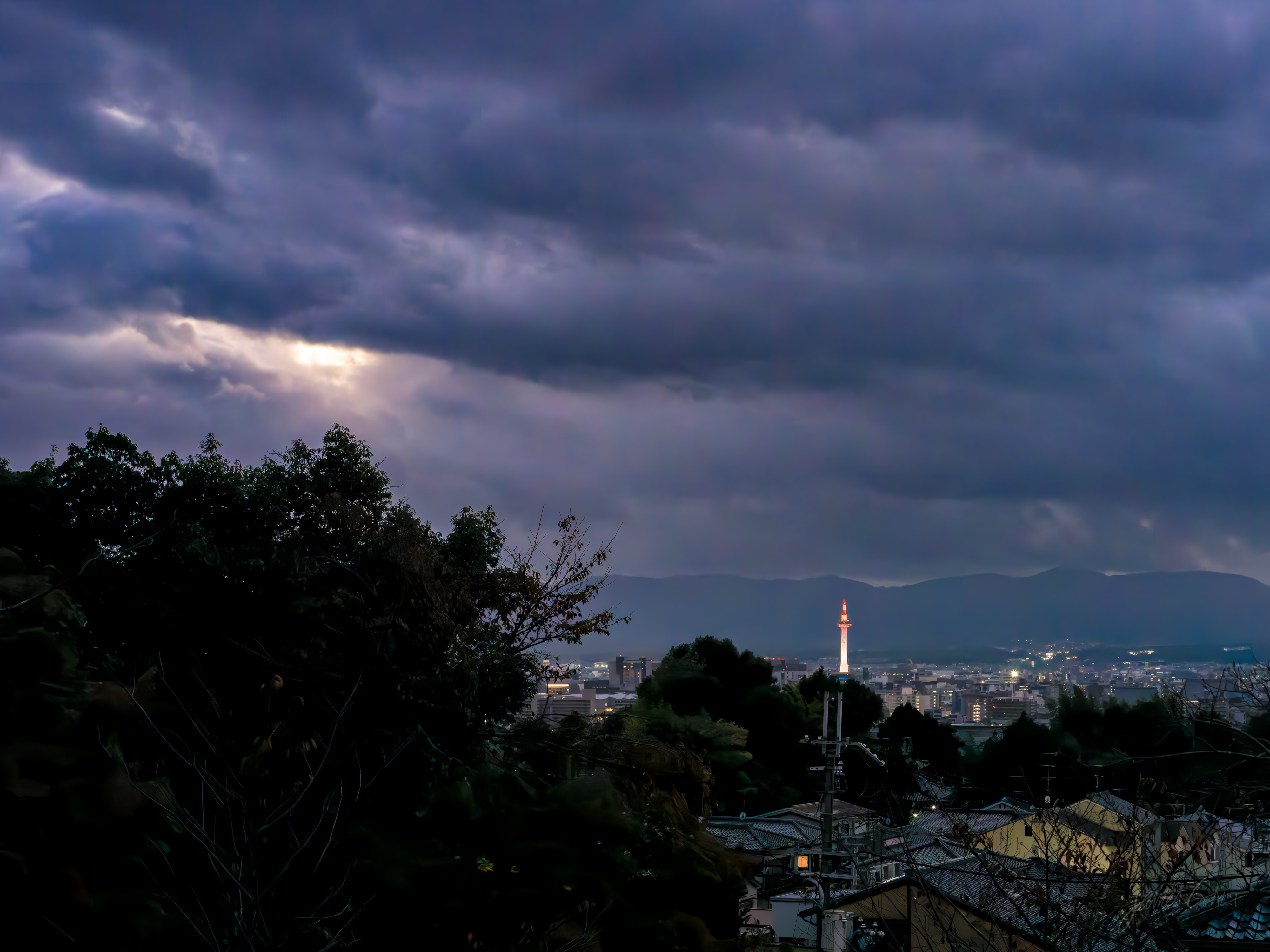 Dusk over Kyoto seen from a hillside, heavy purple-grey cloud filling the sky with a faint break of light, the city spread below with lights coming on and the lit Kyoto Tower standing out on the skyline, dark rooftops and trees in the foreground.