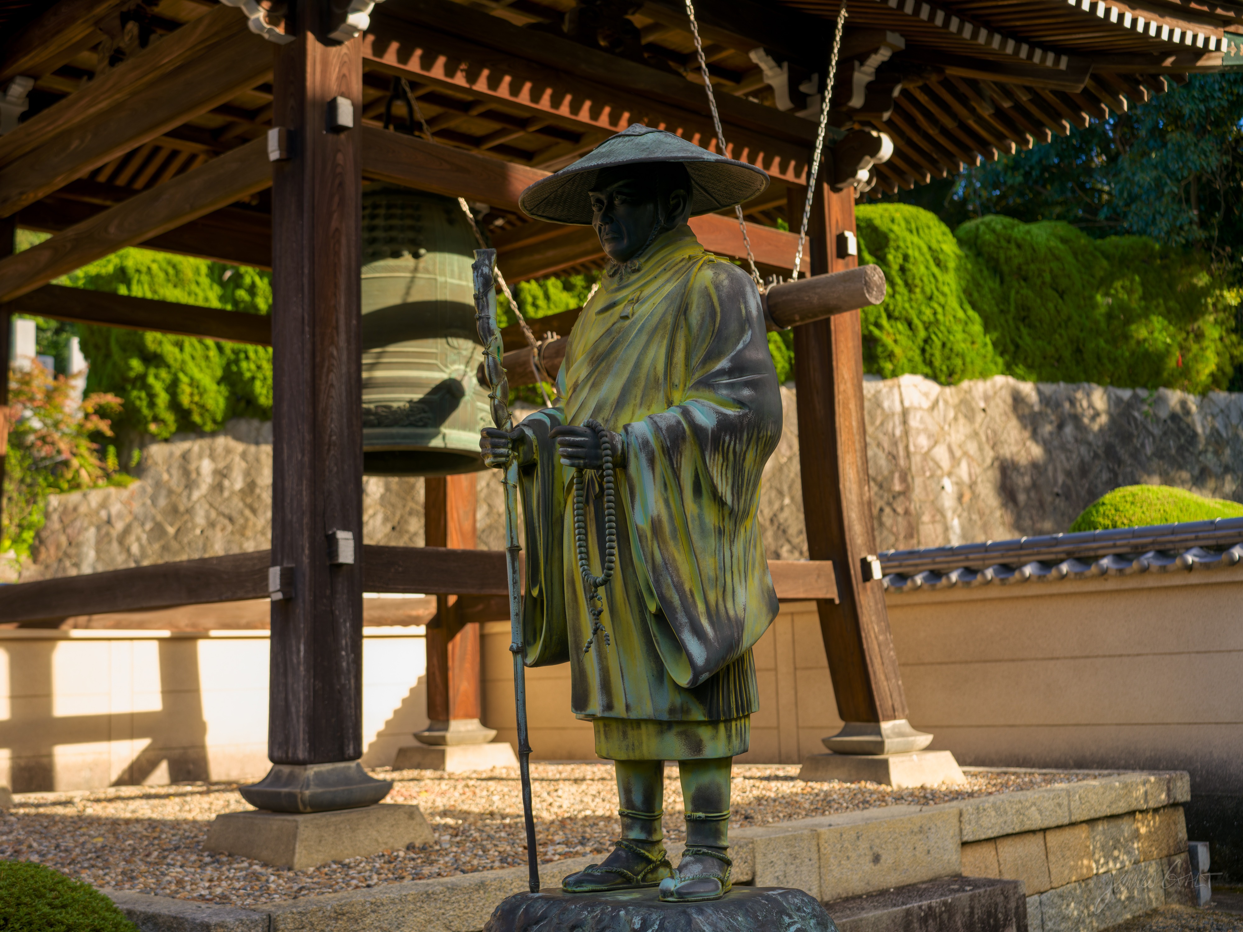 A weathered bronze statue of a traveling monk in a wide hat, holding a staff and prayer beads, standing beside a large temple bell under a wooden bell-house, lit warm by low afternoon sun, clipped shrubs behind.