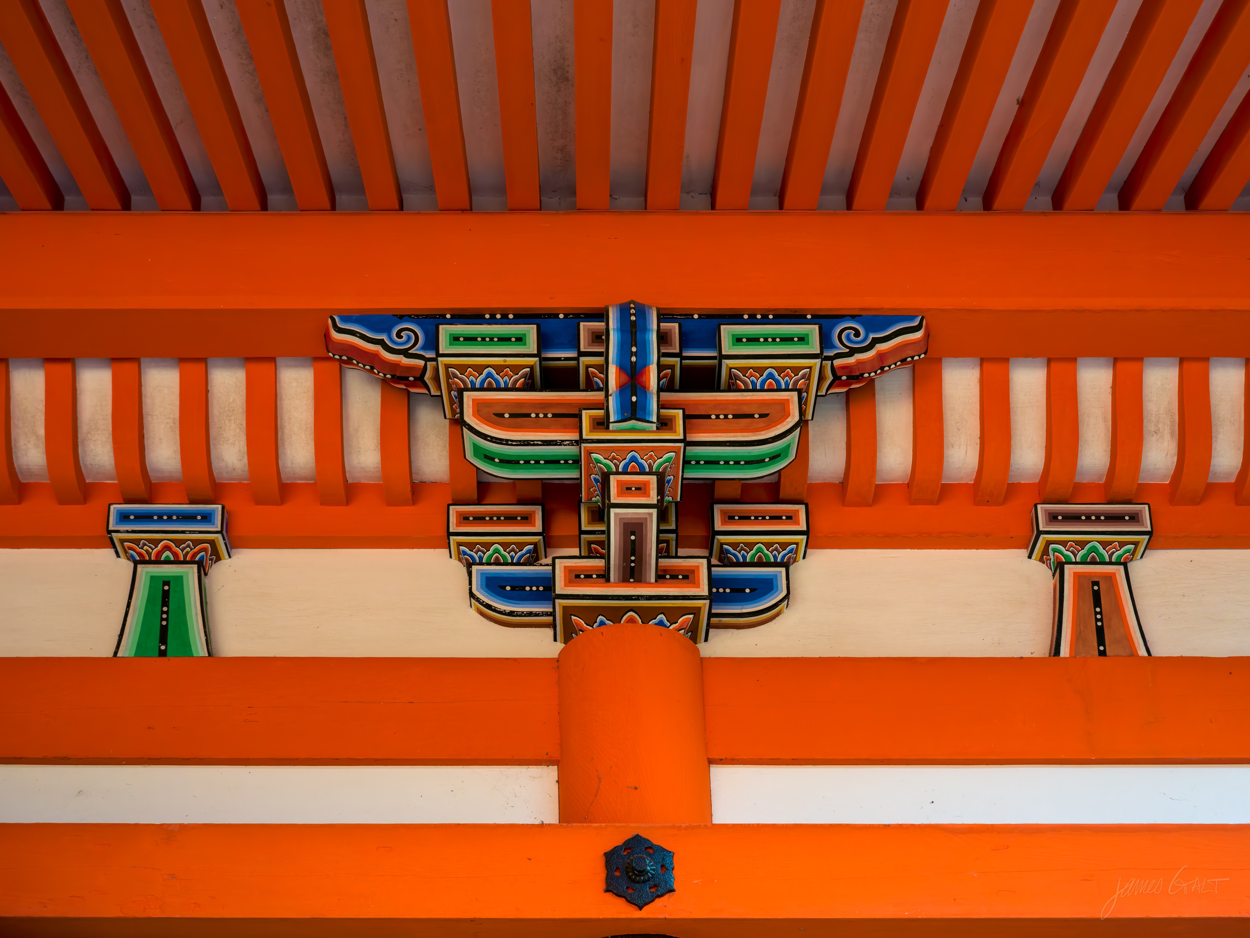 A close-up of a brightly painted temple bracket under the eaves: a symmetrical arrangement of vermilion beams with bracket arms picked out in blue, green, orange and gold, a metal boss at the centre of the lowest beam.