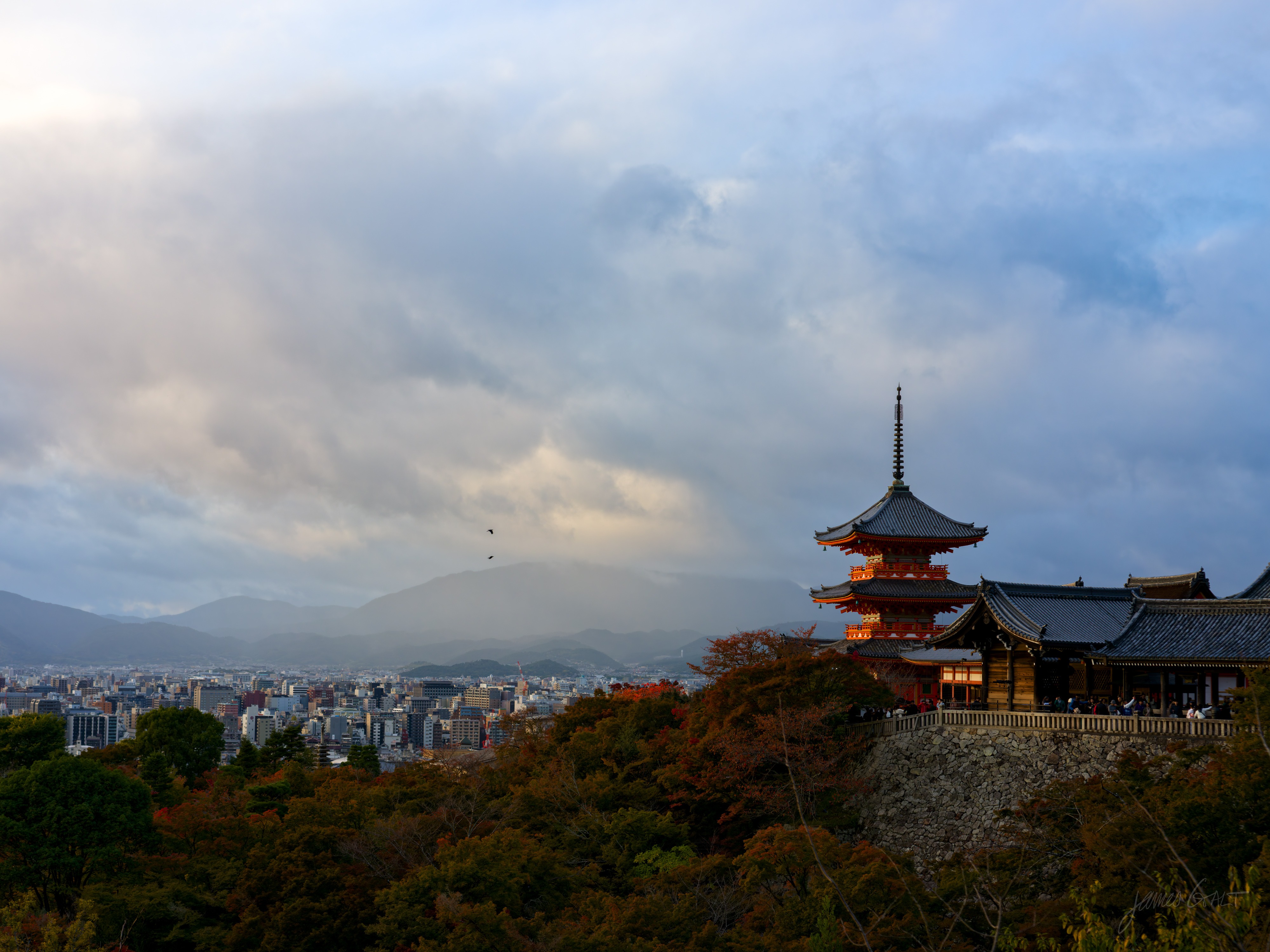 A three-storey vermilion pagoda and temple hall standing on a wooded ridge above the city of Kyoto at golden hour, the buildings and a tall crane on the plain below, a mountain range and dramatic cloud behind, a single bird in the sky.