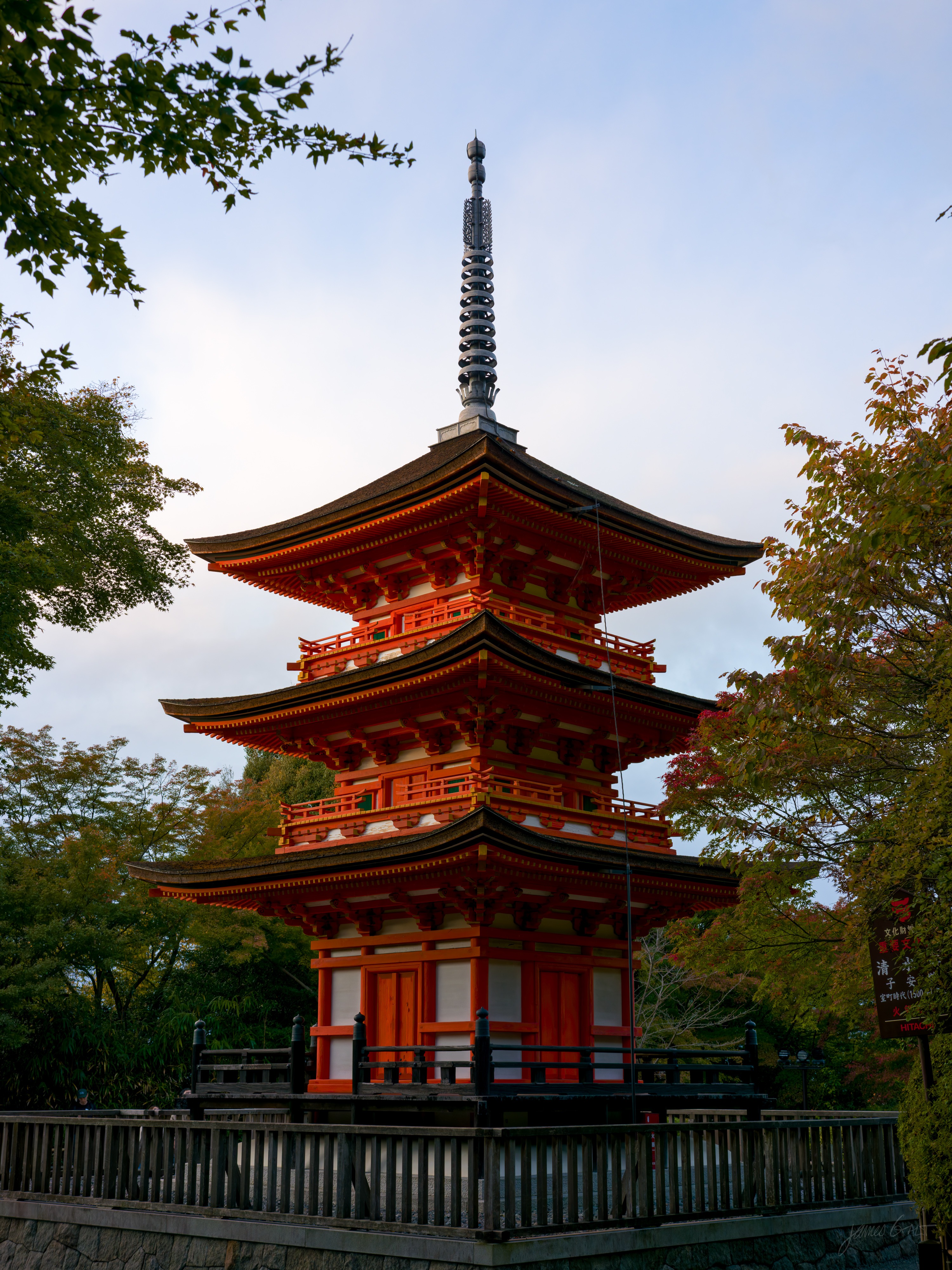 A three-storey vermilion pagoda with dark tiled roofs and a tall finial, framed by green and lightly turning maples against a pale evening sky, a wooden railing around its base.
