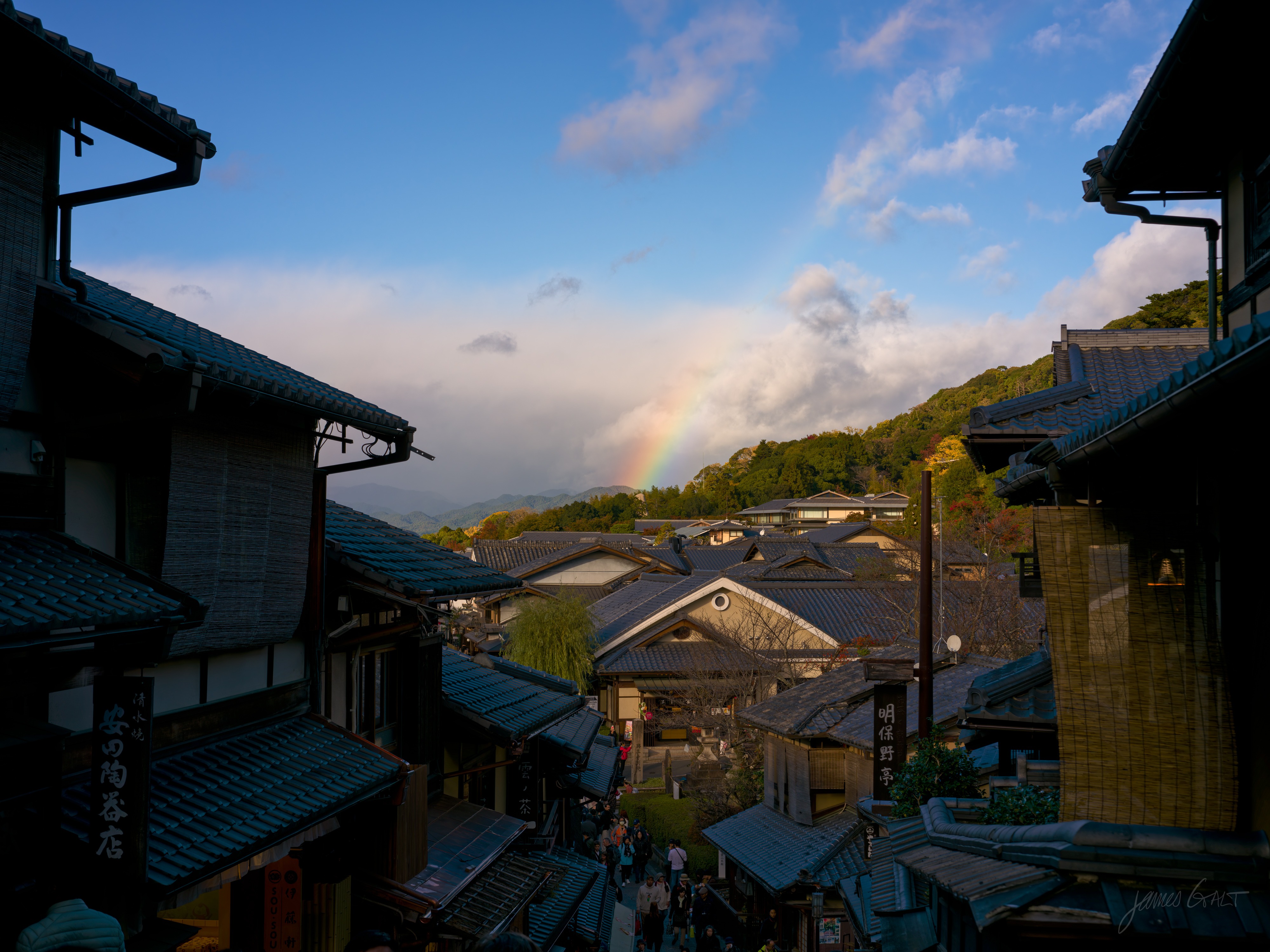 A view down a sloping Higashiyama lane of tiled wooden shopfronts toward wooded hills, a faint rainbow arcing down into the trees under a blue sky with cloud. People walk the street far below, shop signs in Japanese on the buildings.