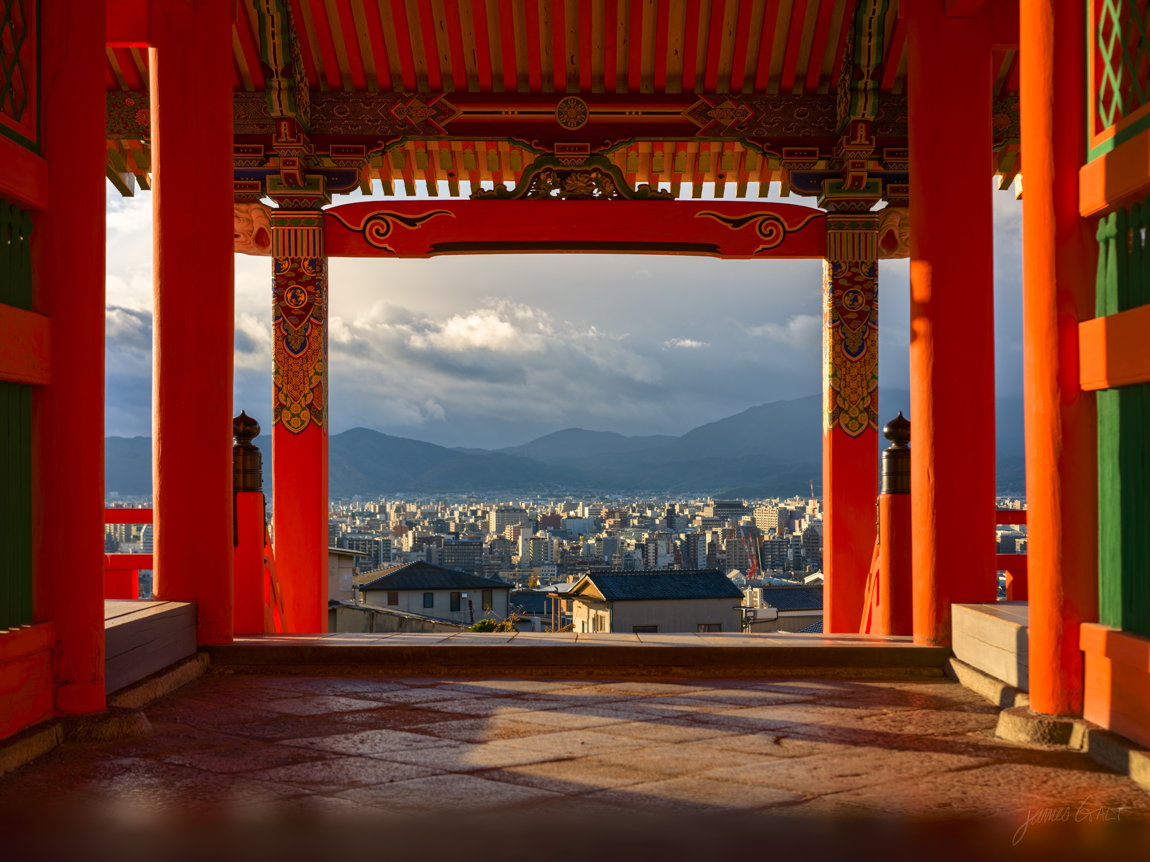 The city of Kyoto framed through the thick vermilion pillars and painted beams of a temple structure, the polished stone floor catching warm light, low mountains and a cloudy sky beyond the buildings.