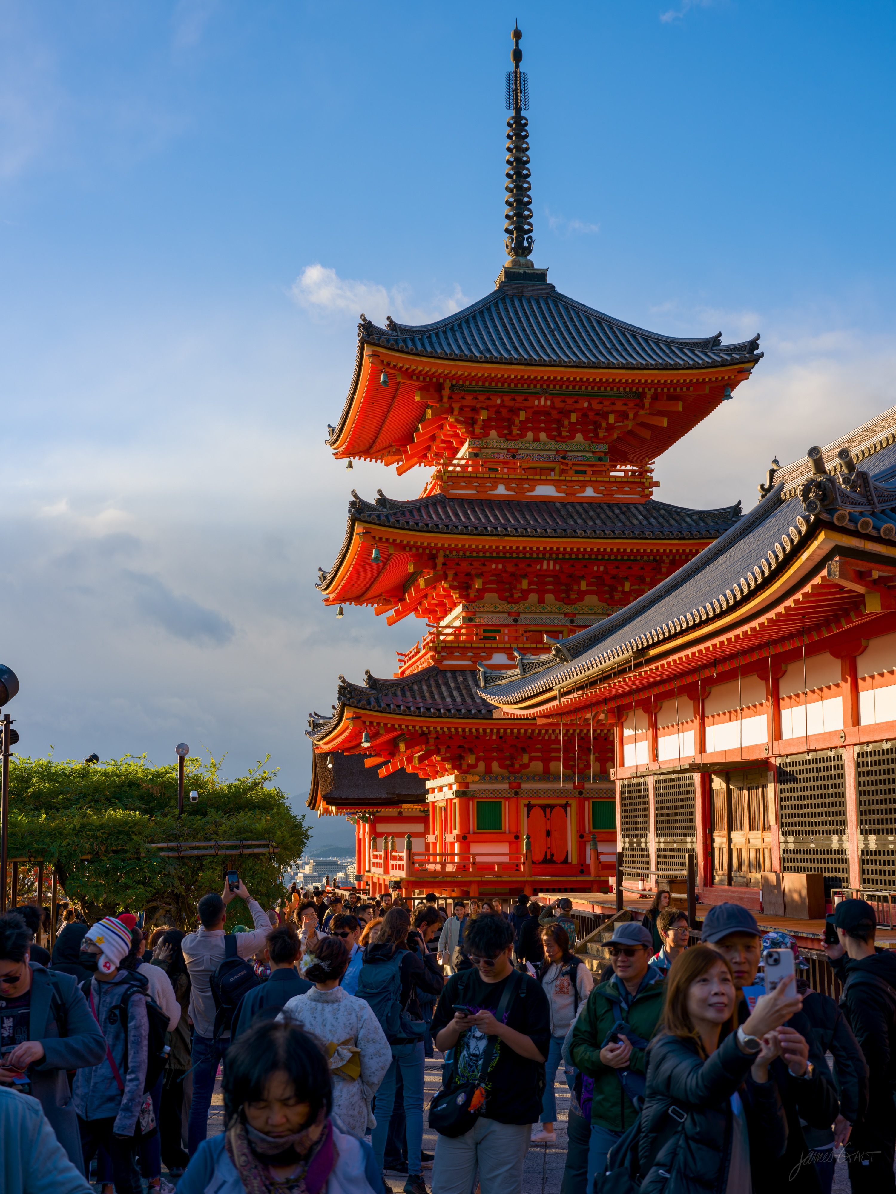 Kiyomizu-dera's three-storey vermilion pagoda and main hall rising against a blue sky in golden light, with a dense crowd of visitors filling the foreground, several holding up phones.