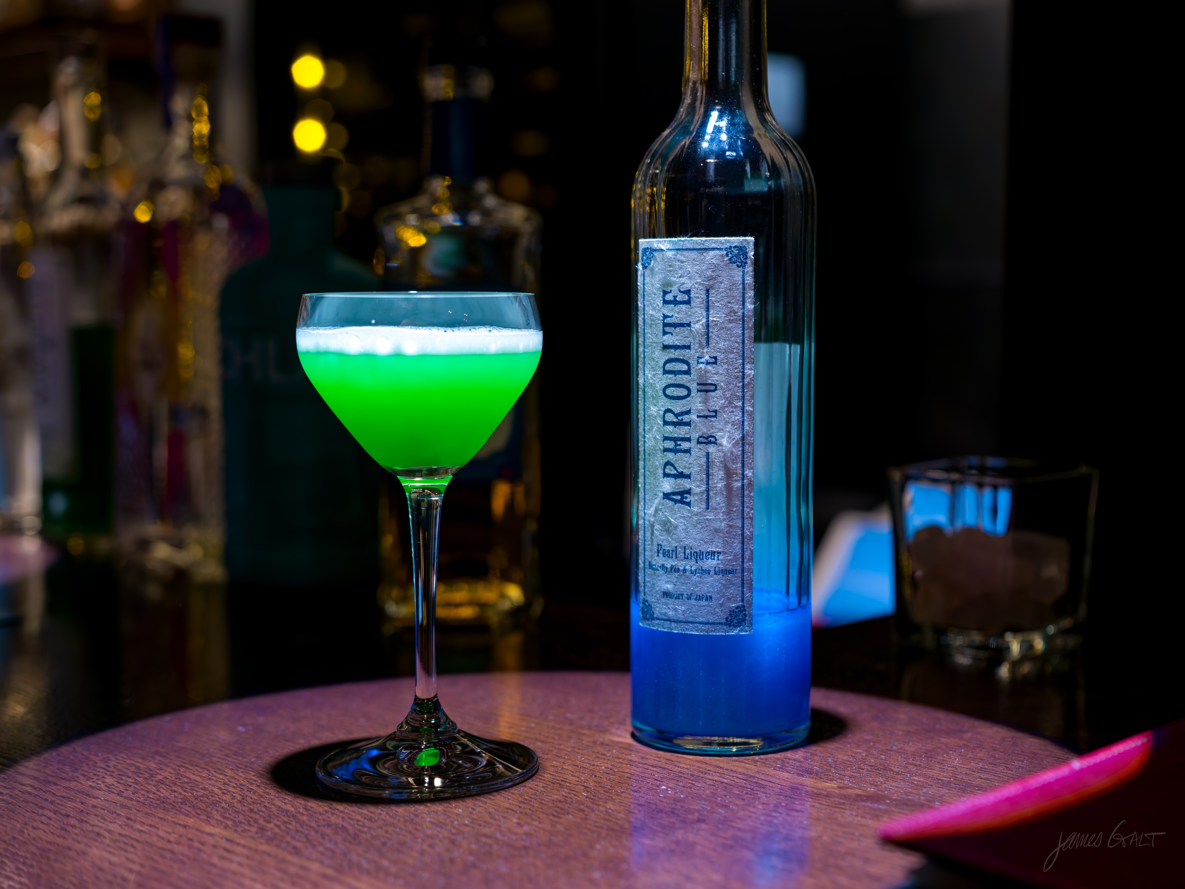 A coupe glass of vivid green cocktail with a frothy top sits on a dark wooden bar beside a tall bottle of Japanese liqueur labelled Aphrodite Blue Pearl Liqueur, its contents shading from clear to deep blue at the base. Out-of-focus bottles and an empty tumbler sit behind under warm bar light.
