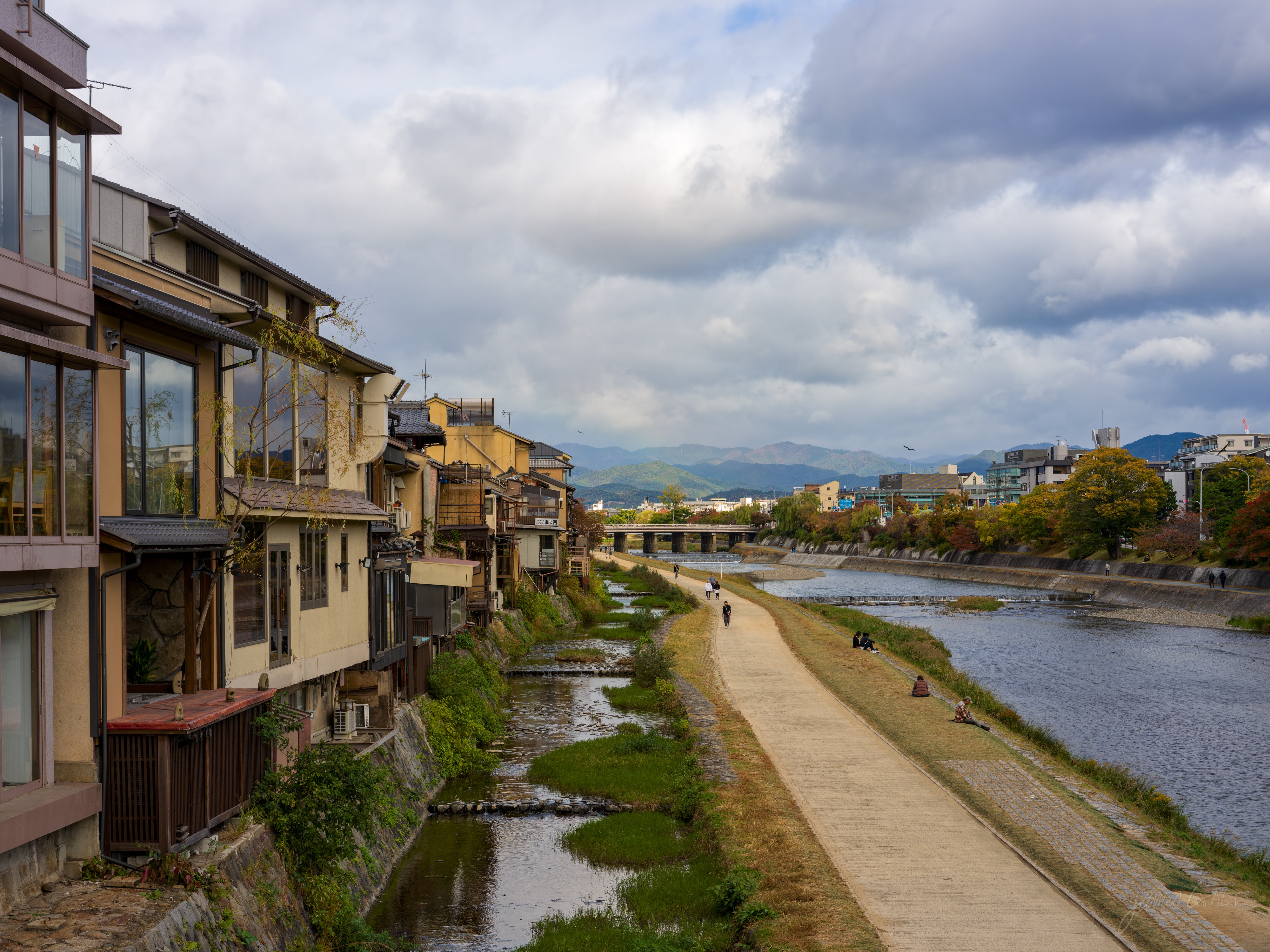 A wide view along the Kamo River in the morning, traditional wooden machiya houses lining the left bank above a low stone embankment, a riverside path on the right with a few walkers, and tree-covered hills in the distance under a cloudy sky.