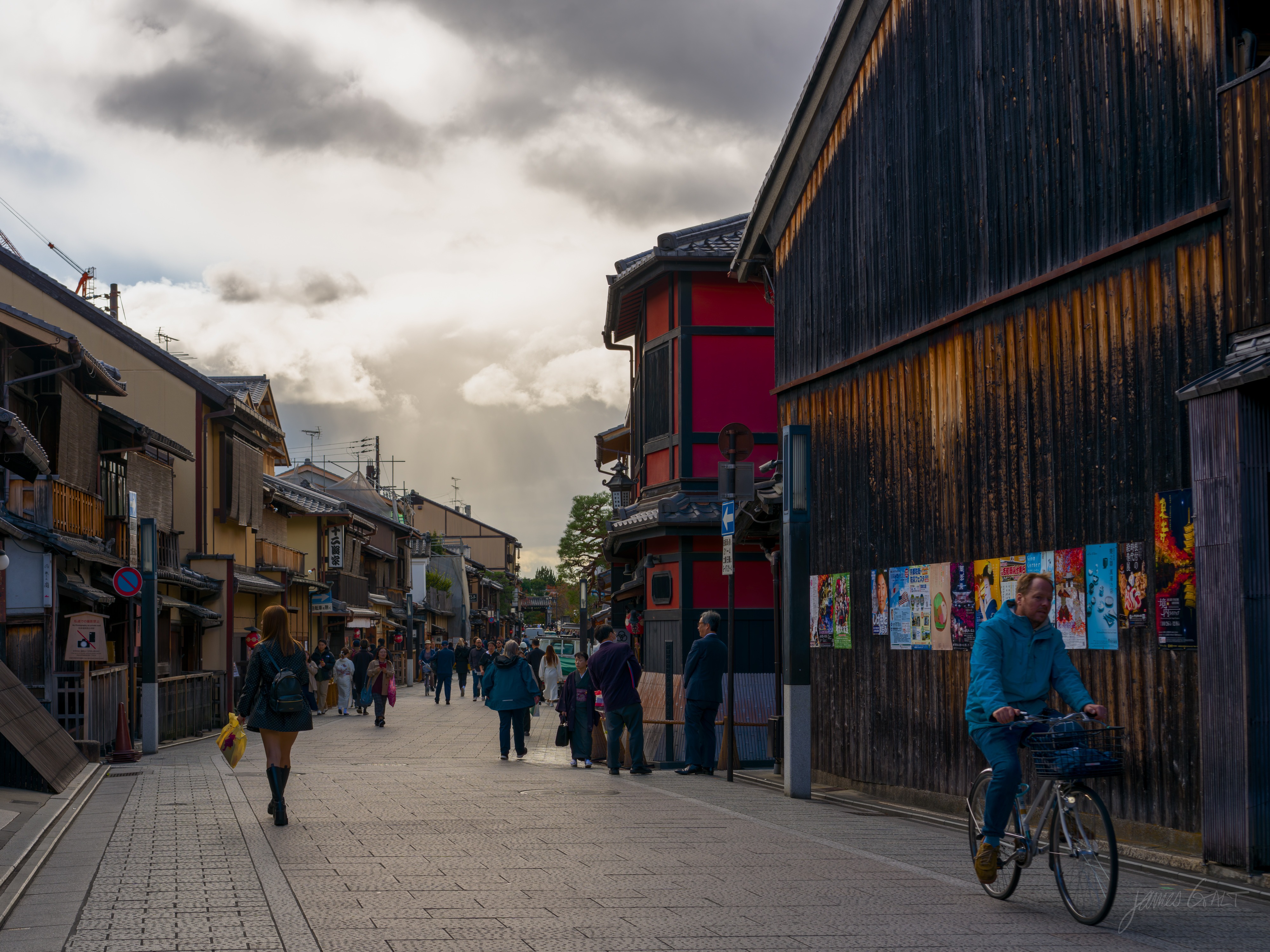 A Gion street in soft light, lined with wooden machiya, a dark-timber teahouse on the right and a red-walled building beyond. People walk the lane and a man in a blue jacket rides a bicycle toward the camera, dramatic clouds above.