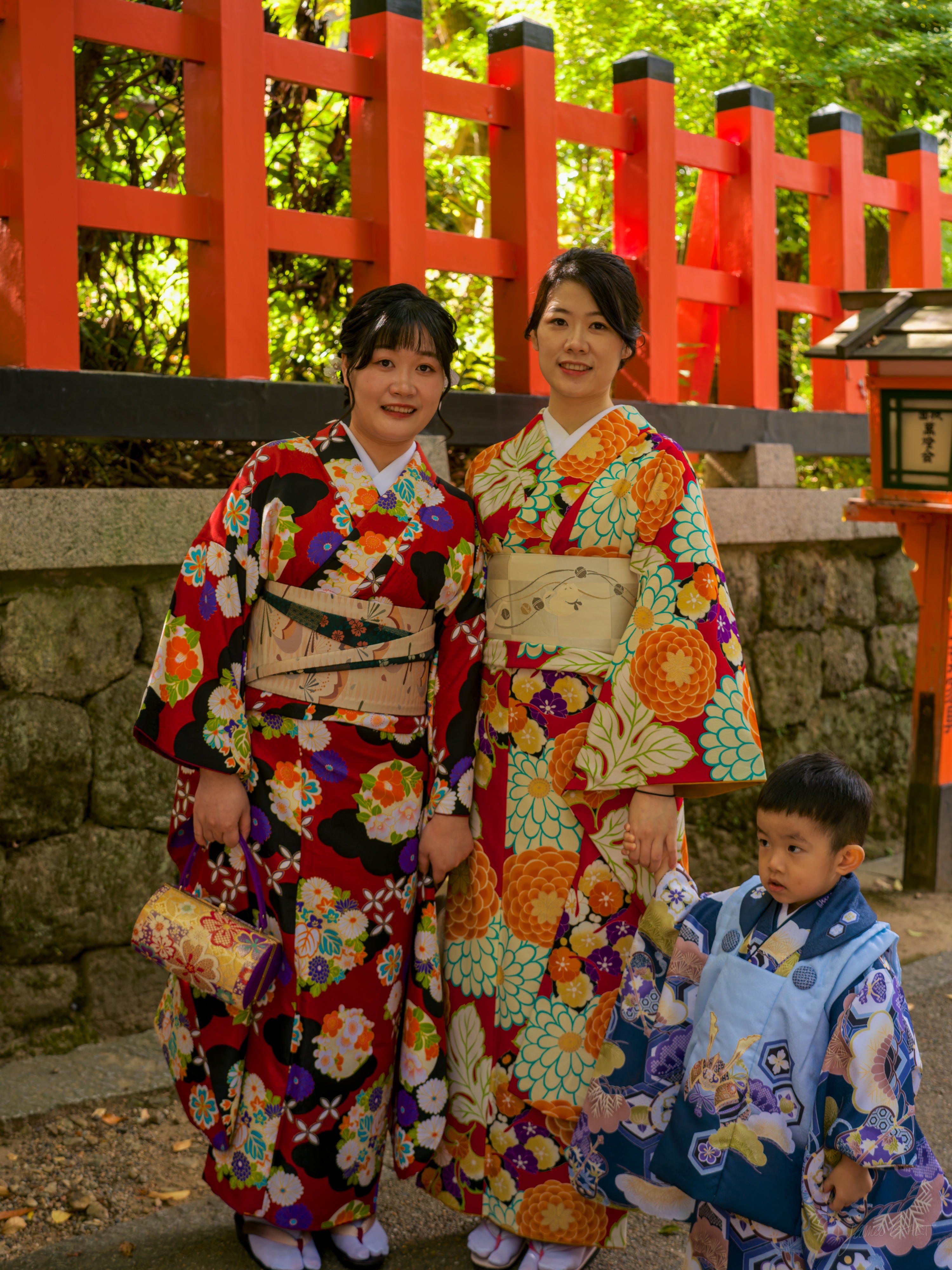 Two women in vivid floral kimono, deep red and orange, stand smiling for the camera beside a vermilion fence, a small boy in a pale blue patterned kimono standing shyly to their right. Green foliage behind.