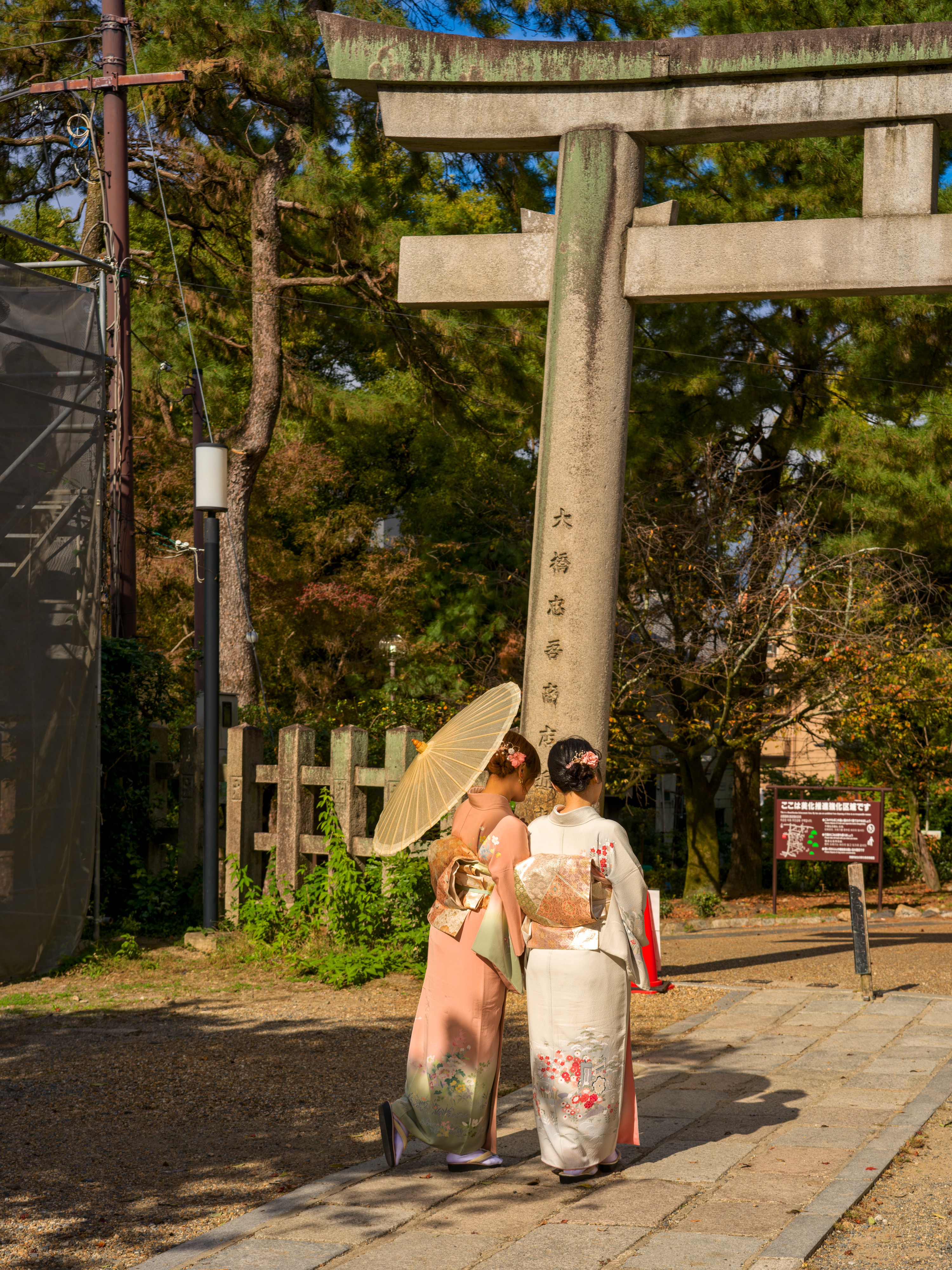 Two women in kimono seen from behind, one in pale pink holding up a parasol, the other in cream with a red-lined hem, walking toward a tall stone torii in golden morning light, an inscription carved down the torii's pillar.