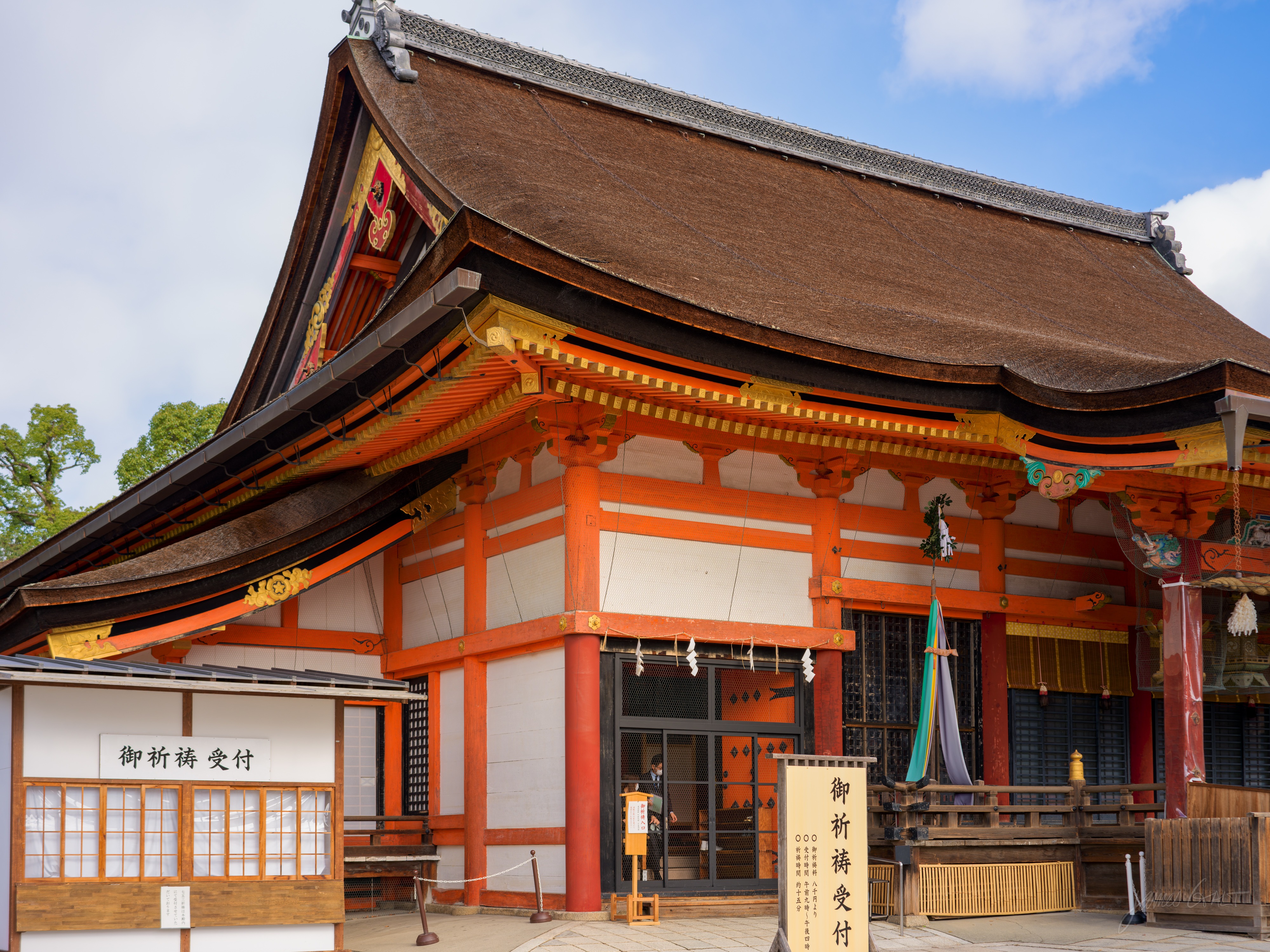 The main hall of Yasaka Shrine, a large vermilion building with white walls, gold and orange trim and a broad sweeping cypress-bark roof. Signs reading prayer reception stand out front, a small figure in the doorway.