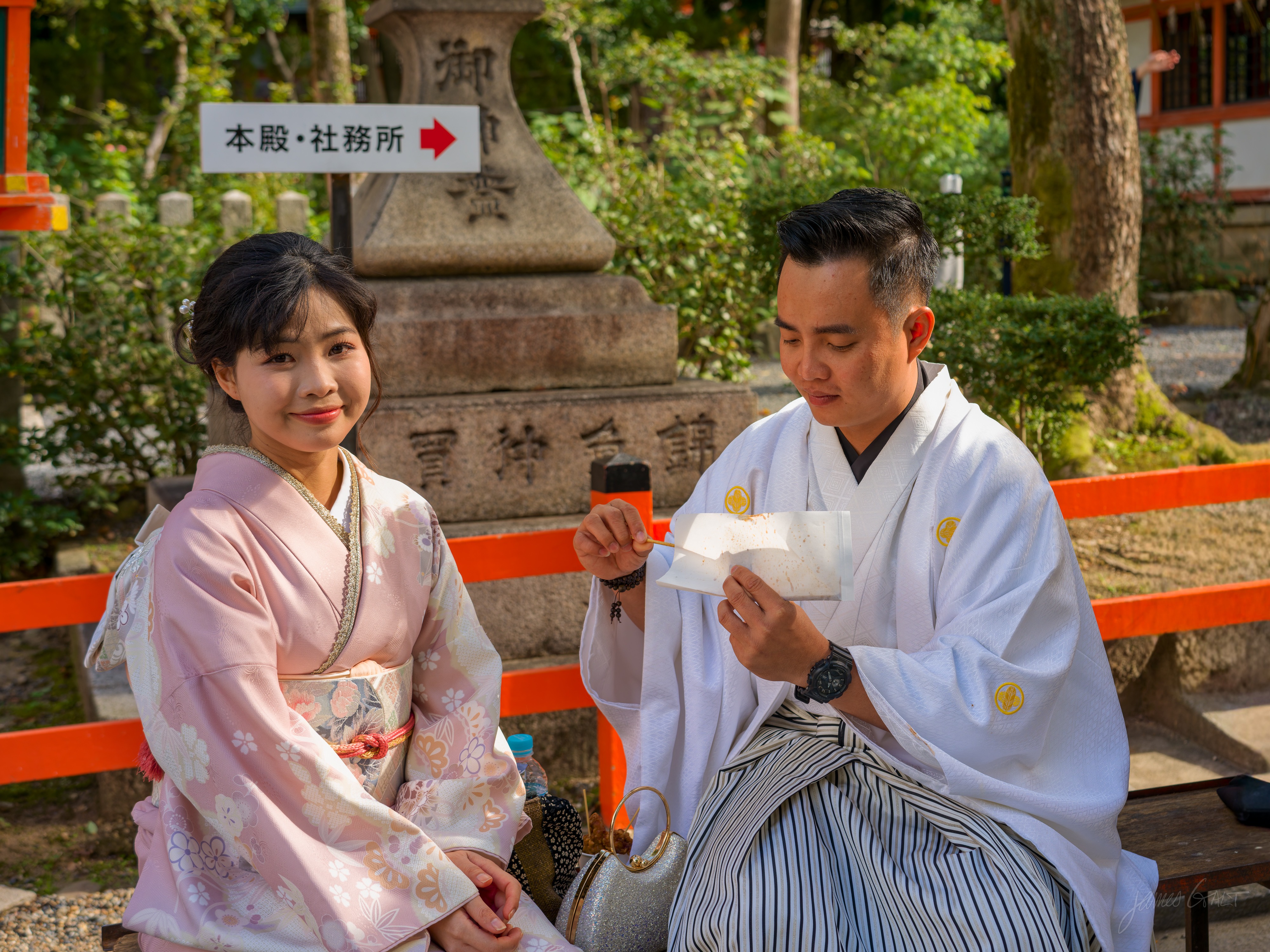 A young couple in formal traditional dress sit on a red bench at the shrine, the woman in a pale pink kimono, the man in a white kimono and striped hakama reading a slip of paper. A stone lantern and a vermilion fence stand behind them.