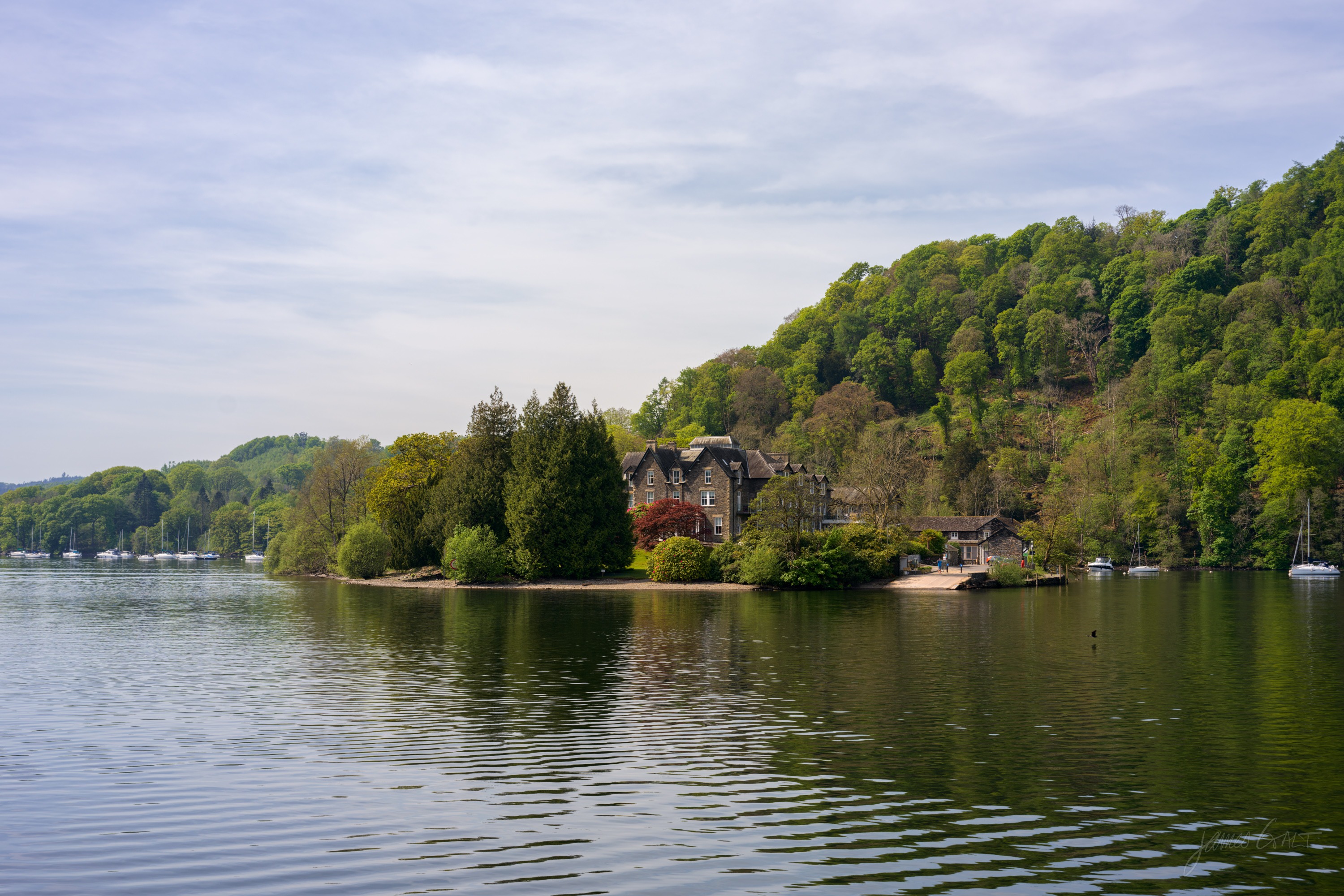 A stone house part-hidden in trees on a wooded promontory jutting into a calm lake, a steep green hillside rising behind, still water and moored yachts in the distance.