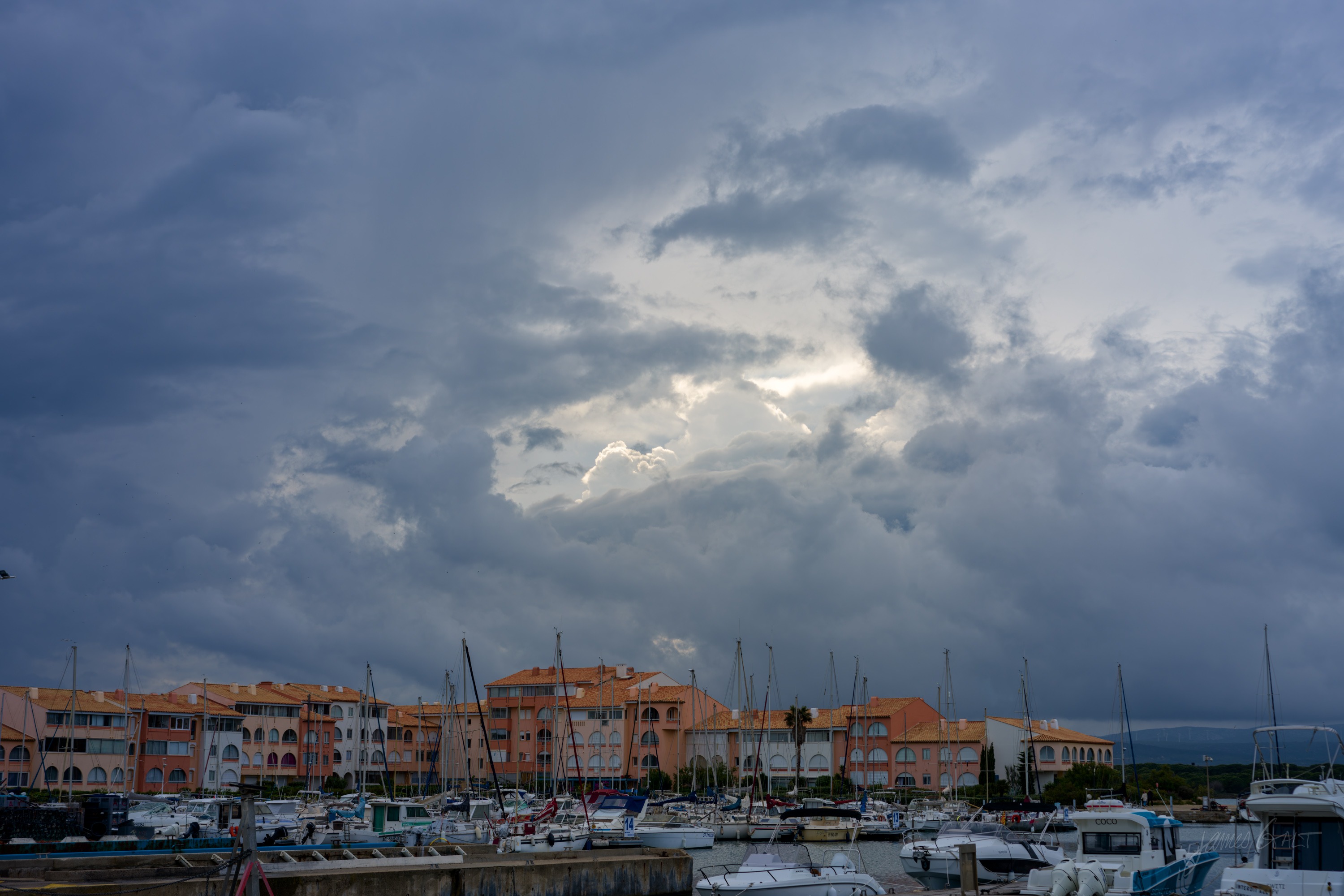A marina of moored yachts in front of low pastel apartment blocks, a heavy bank of storm cloud breaking to a bright gap over the water.