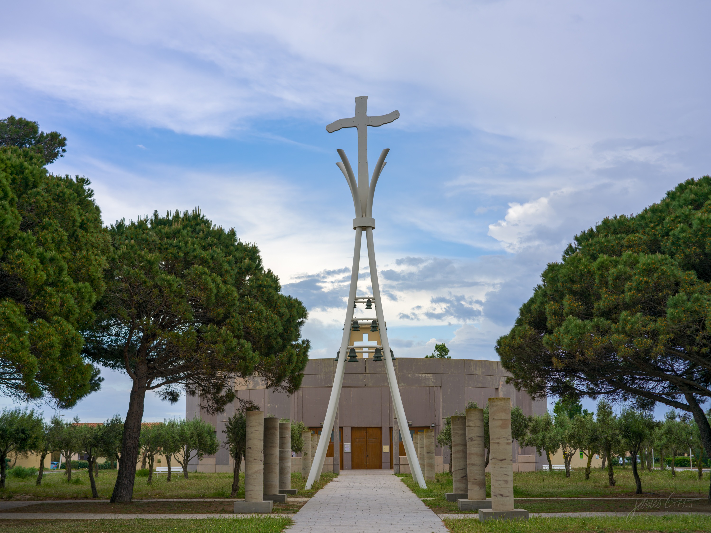 A modern concrete church with a tall white sculpted cross rising in front of it, flanked by umbrella pines, a bright evening sky behind.