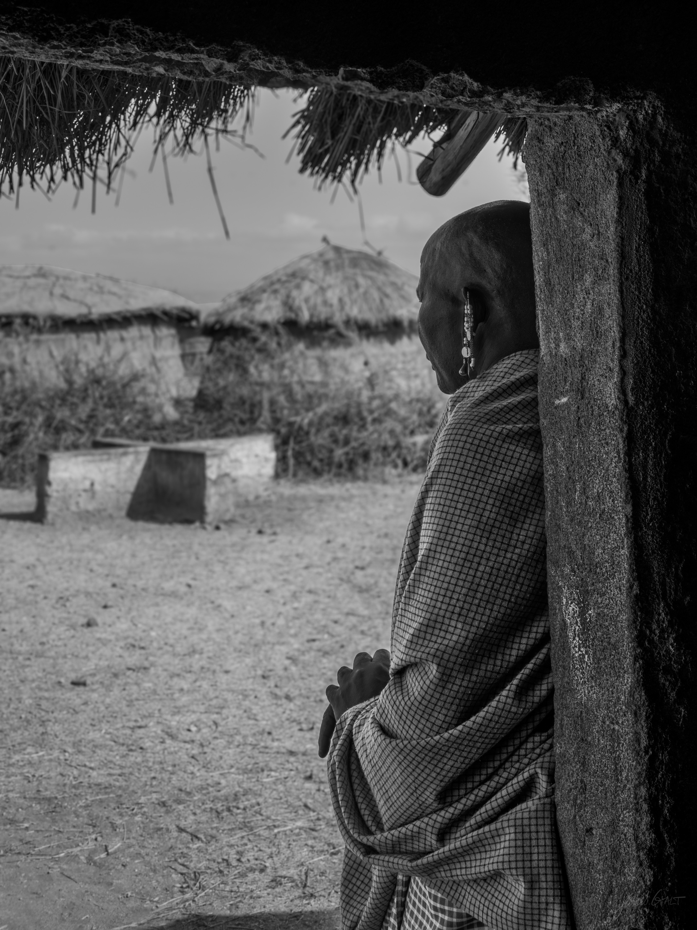 Black and white photograph of a Maasai figure standing in the low doorway of a thatch-roofed house, dark interior behind, huts and dry ground outside.