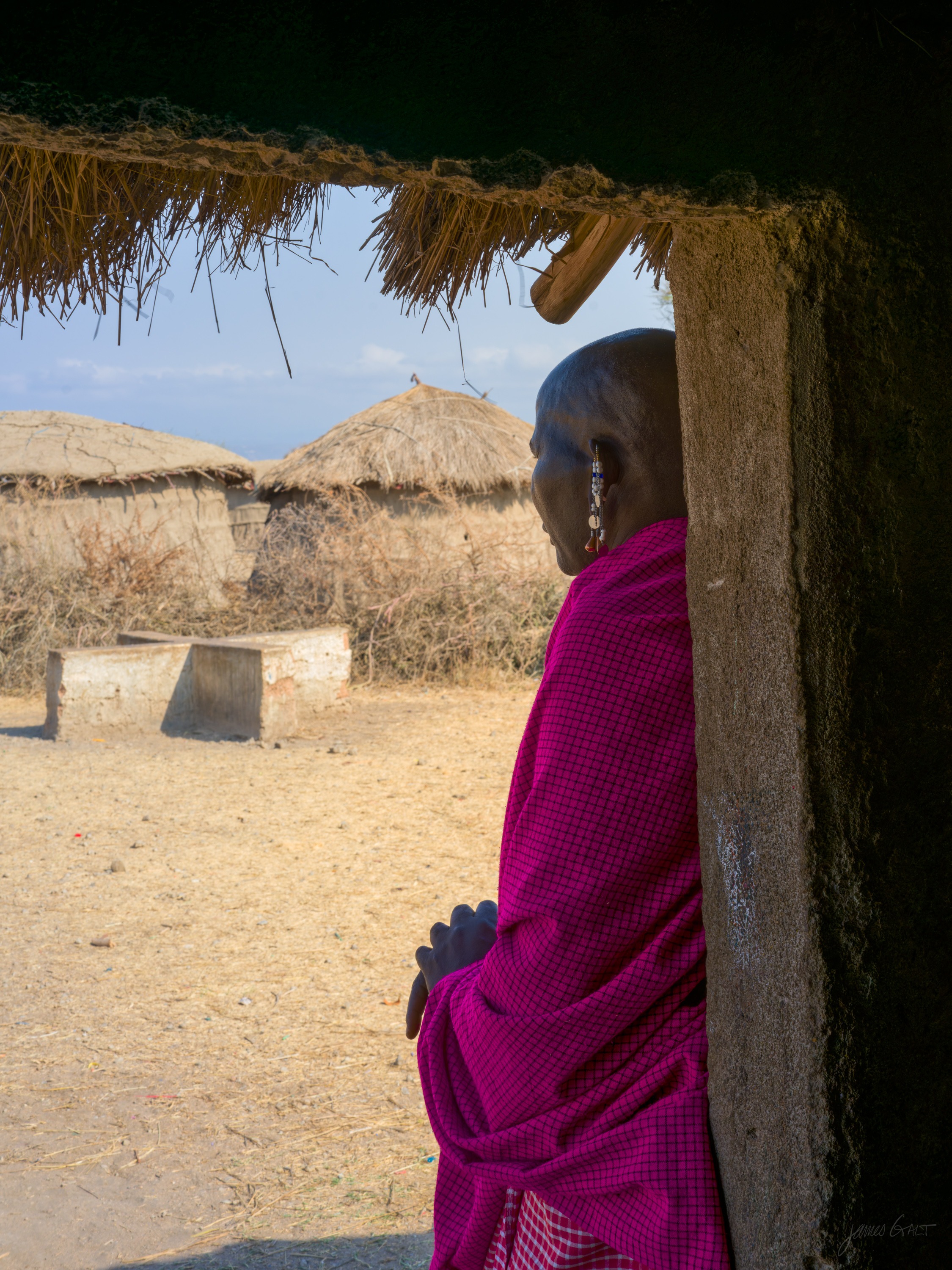 The same doorway in colour, a person in a red shuka standing in the low opening of a mud-walled, thatch-roofed house, the village beyond in daylight.
