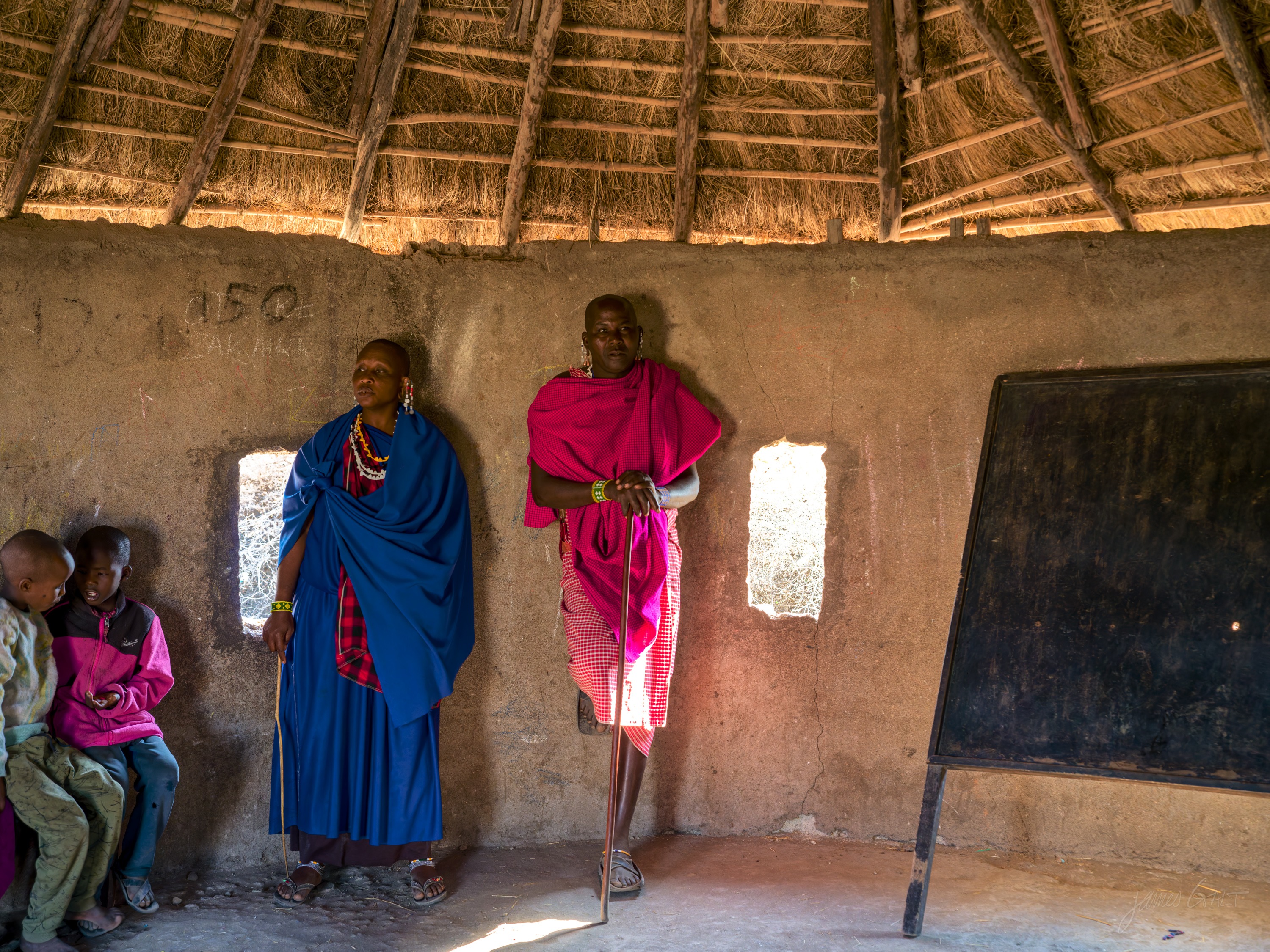The dim interior of a Maasai house in colour, two figures seated and standing against dark walls, a small bright window letting in a shaft of light.