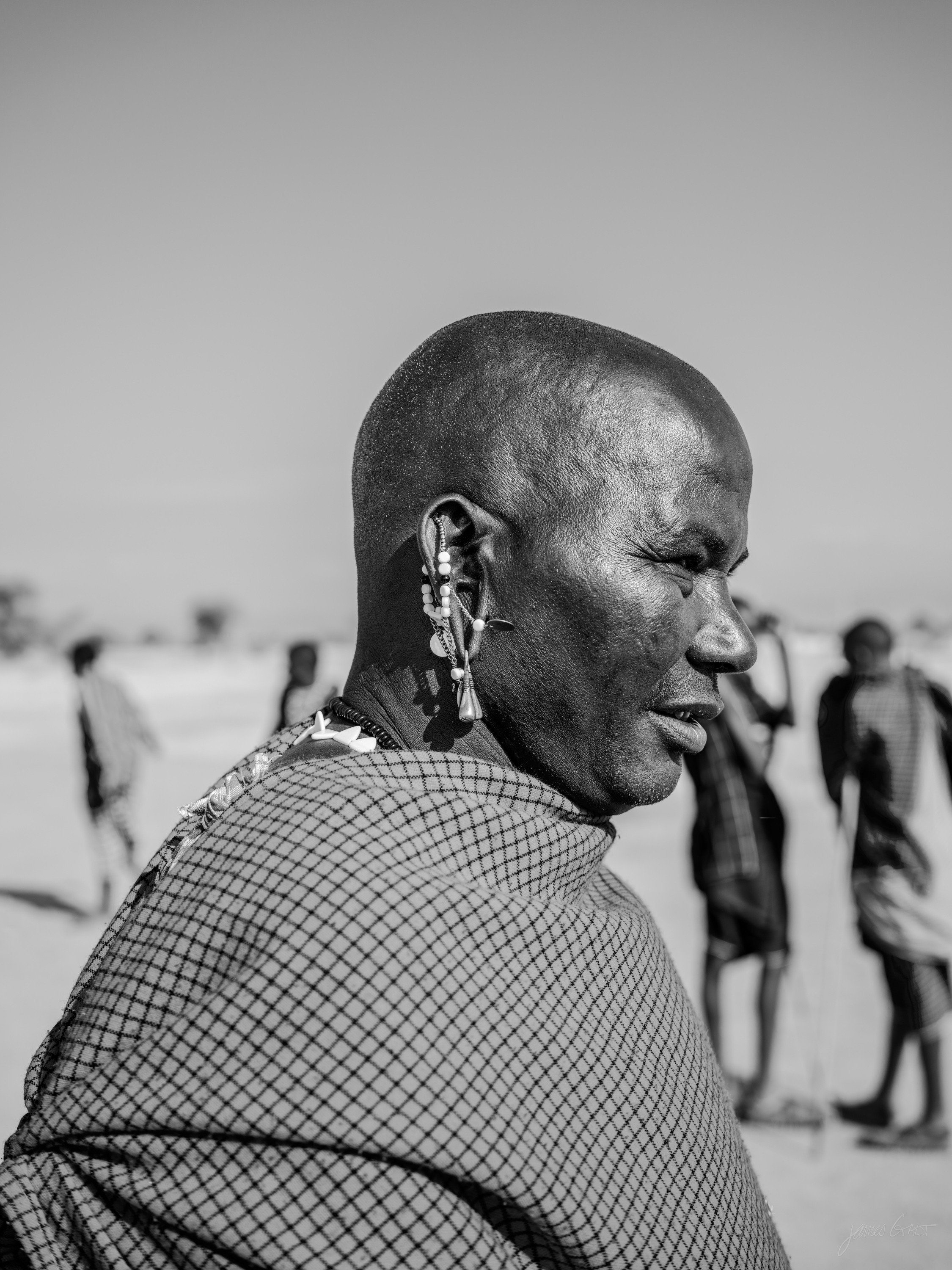 Black and white portrait of a Maasai elder with a lined face and short grey hair, wrapped in a patterned cloth, looking toward the camera.