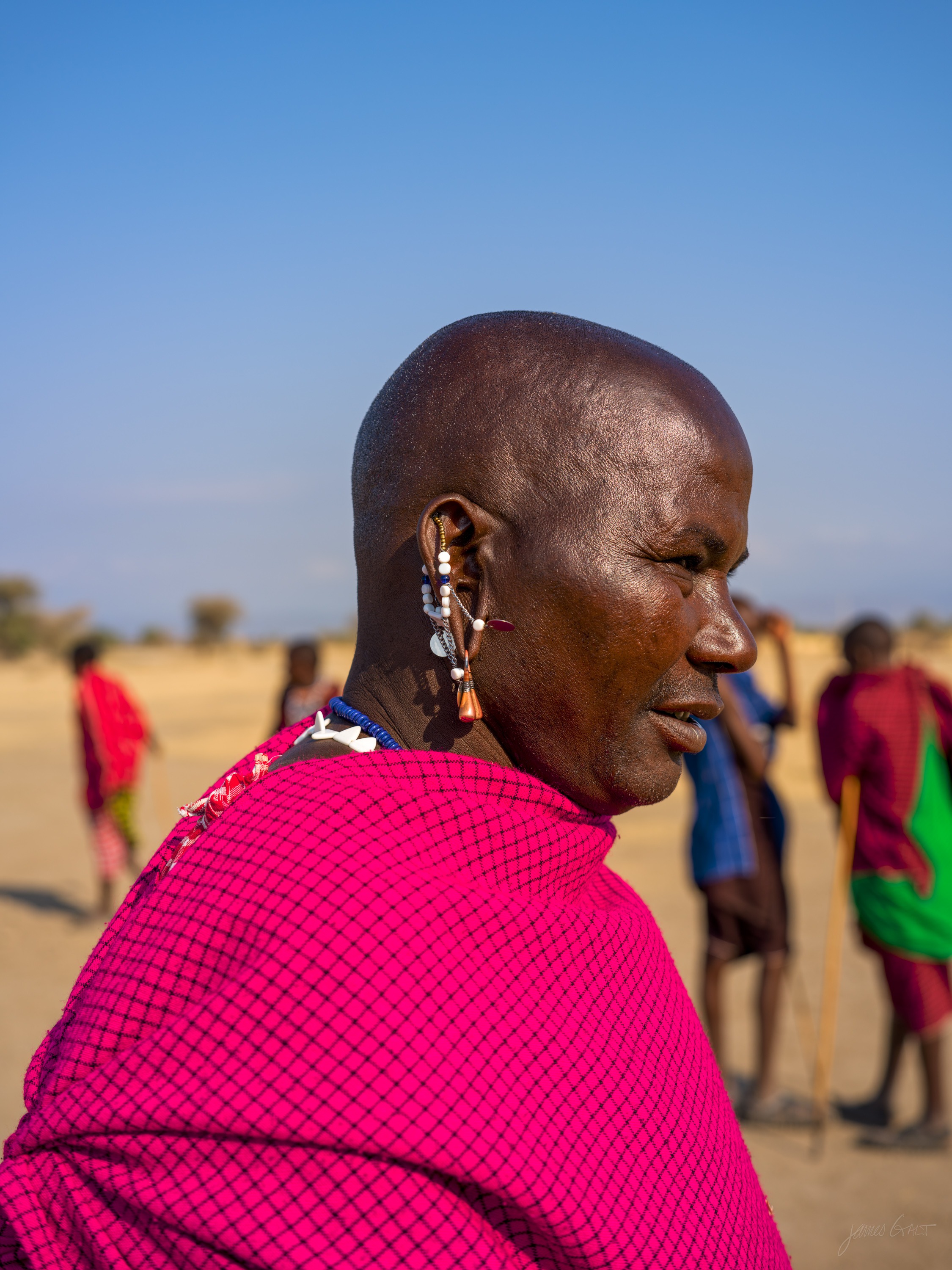 The same elder in colour, in a red and checked robe, standing on dusty ground with other villagers blurred behind him in warm morning light.