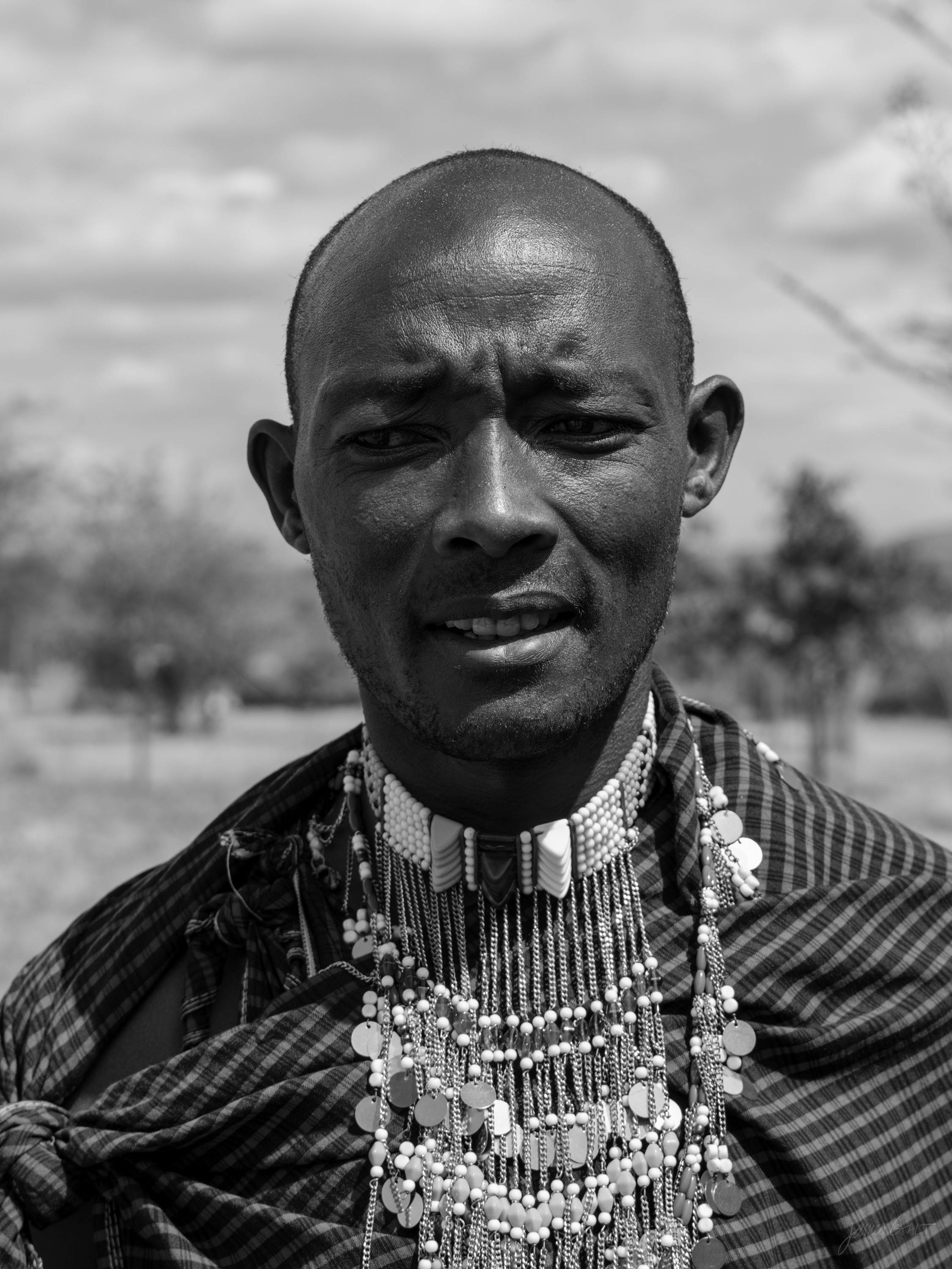 Black and white portrait of a Maasai man with close-cropped hair and a lined face, wearing a striped robe and several beaded necklaces, looking directly at the camera. Blurred acacia savannah stretches behind him.