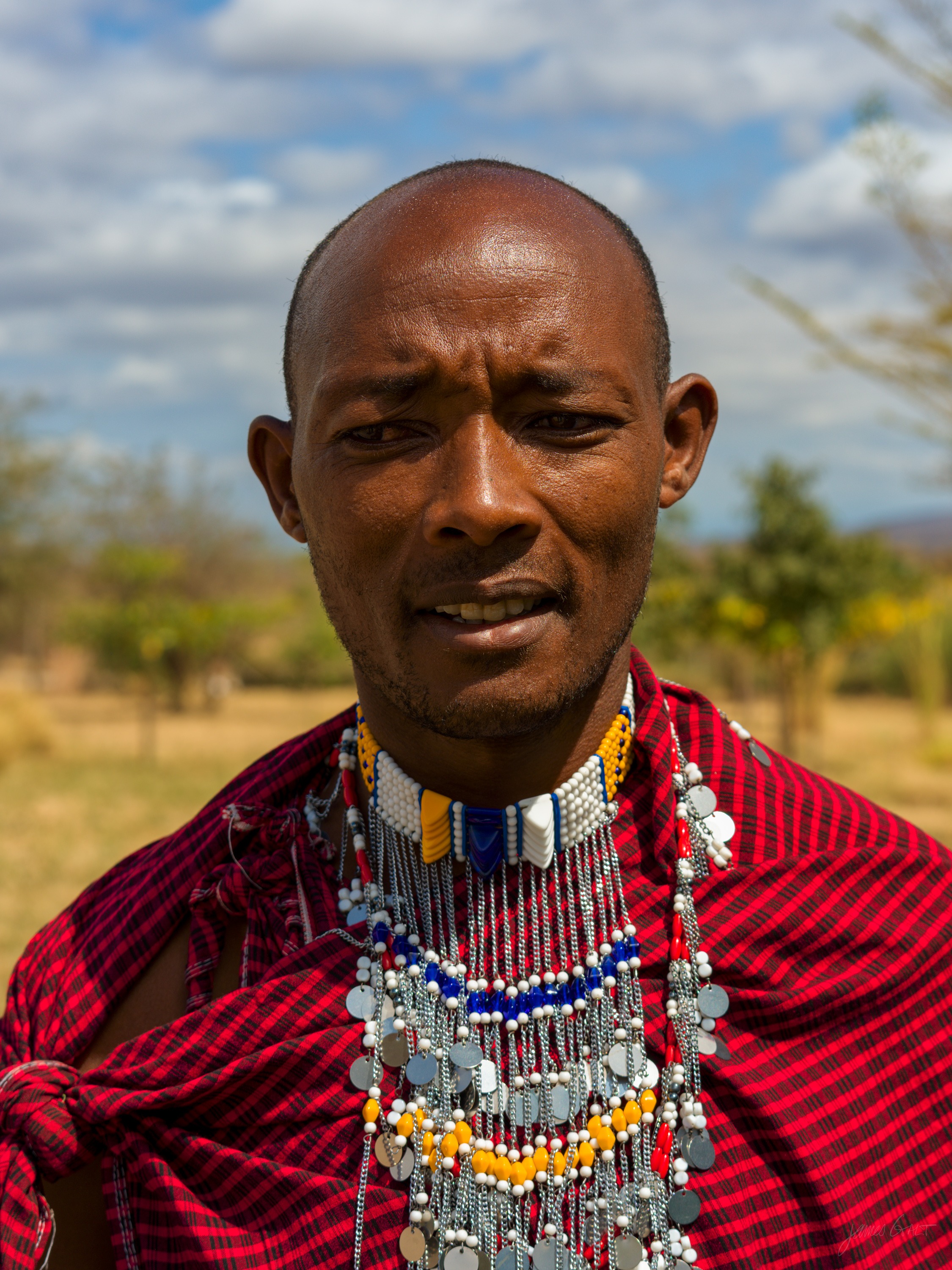 The same man in colour, in a deep red robe with layered white and coloured bead necklaces, the tawny savannah and a hazy mountain behind him under a blue sky.