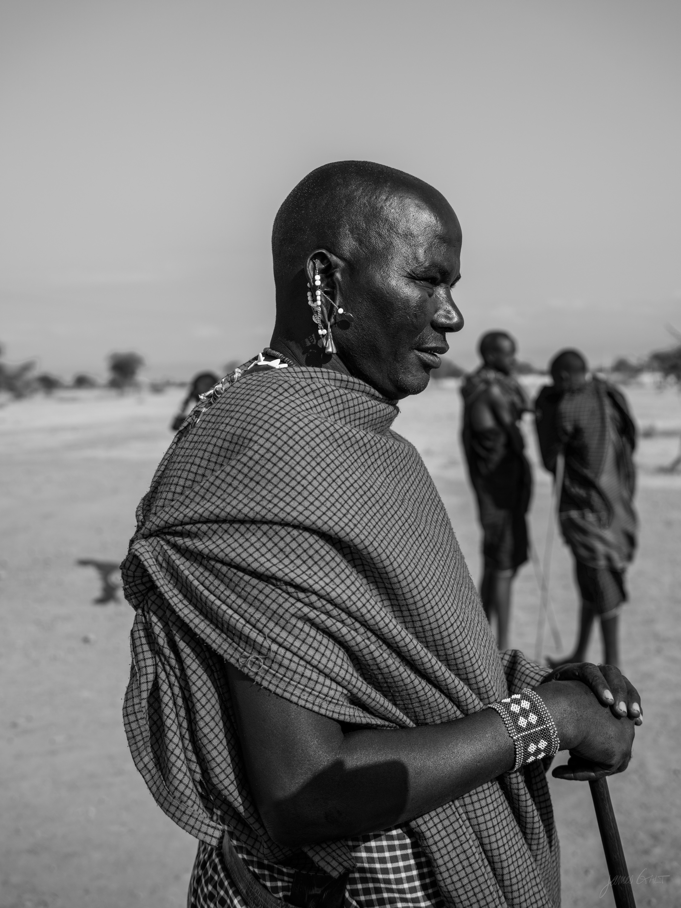 Black and white photograph of a Maasai man standing in profile holding a long staff, wrapped in a patterned shuka, other figures blurred behind him.