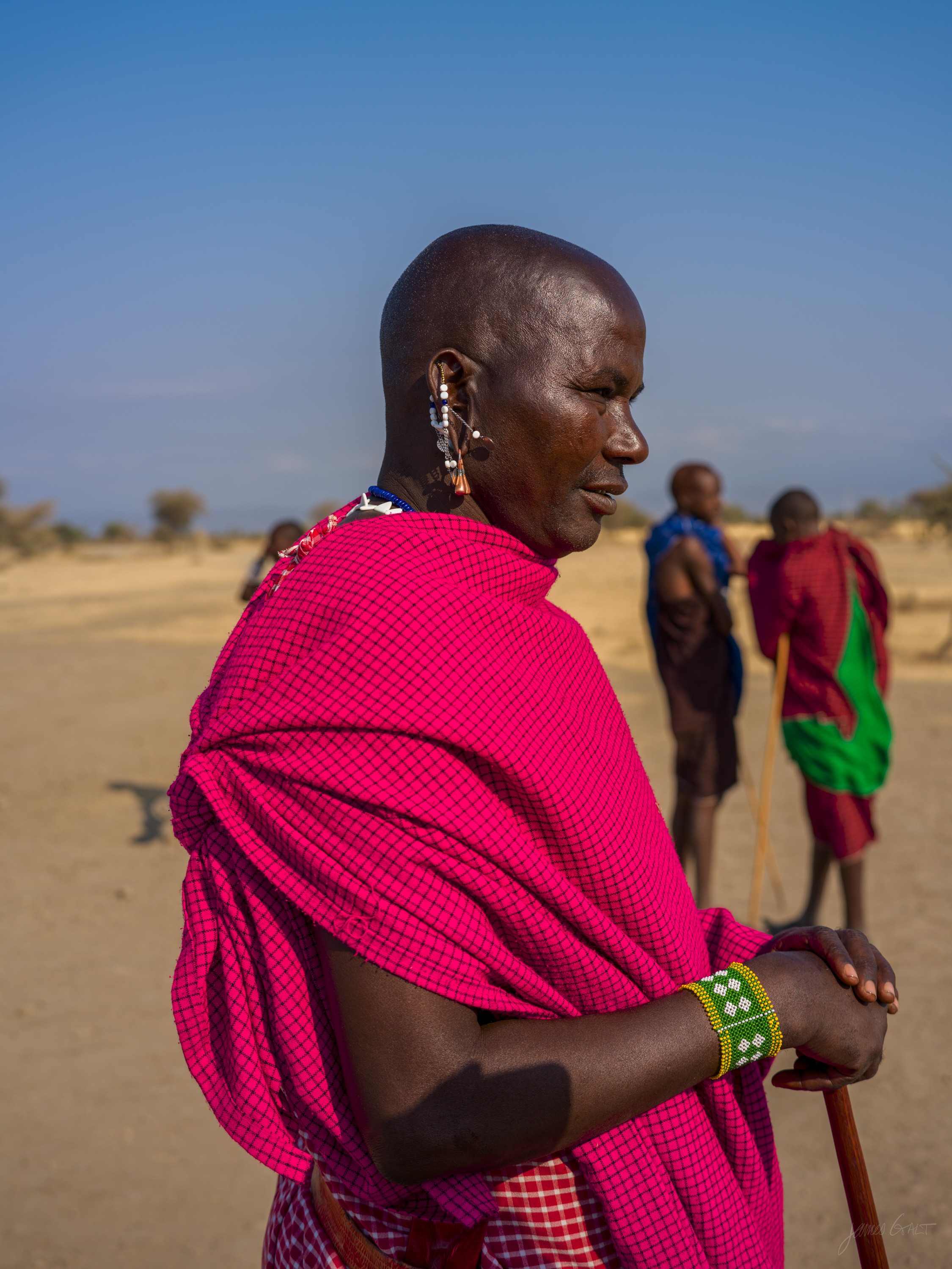 The same man in colour, in a bright pink and red shuka, holding his staff, dusty ground and villagers soft behind him in morning light.