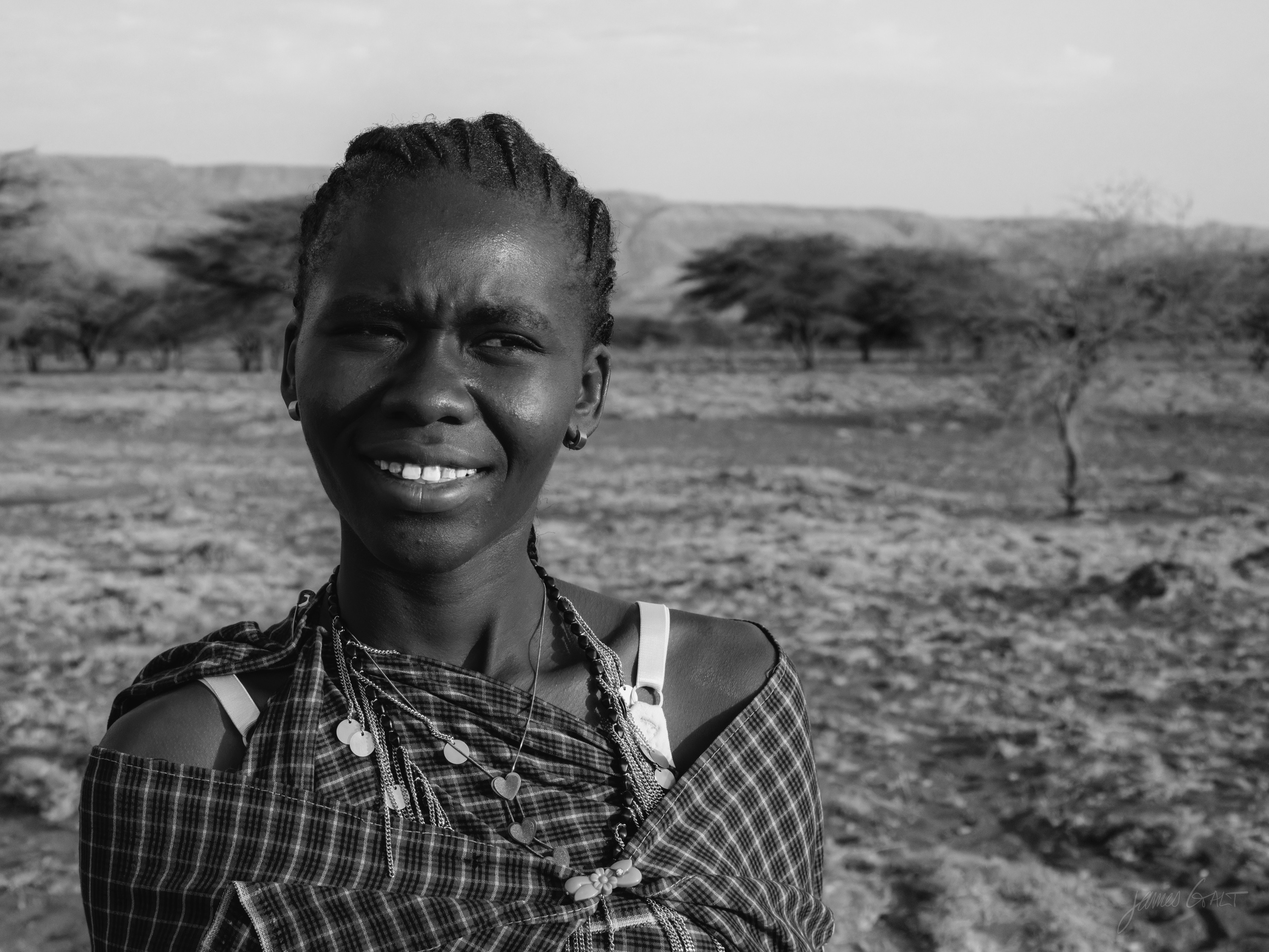 Black and white portrait of a smiling Maasai woman with braided hair and beaded jewellery, warm crinkles at her eyes, acacia savannah soft behind her.