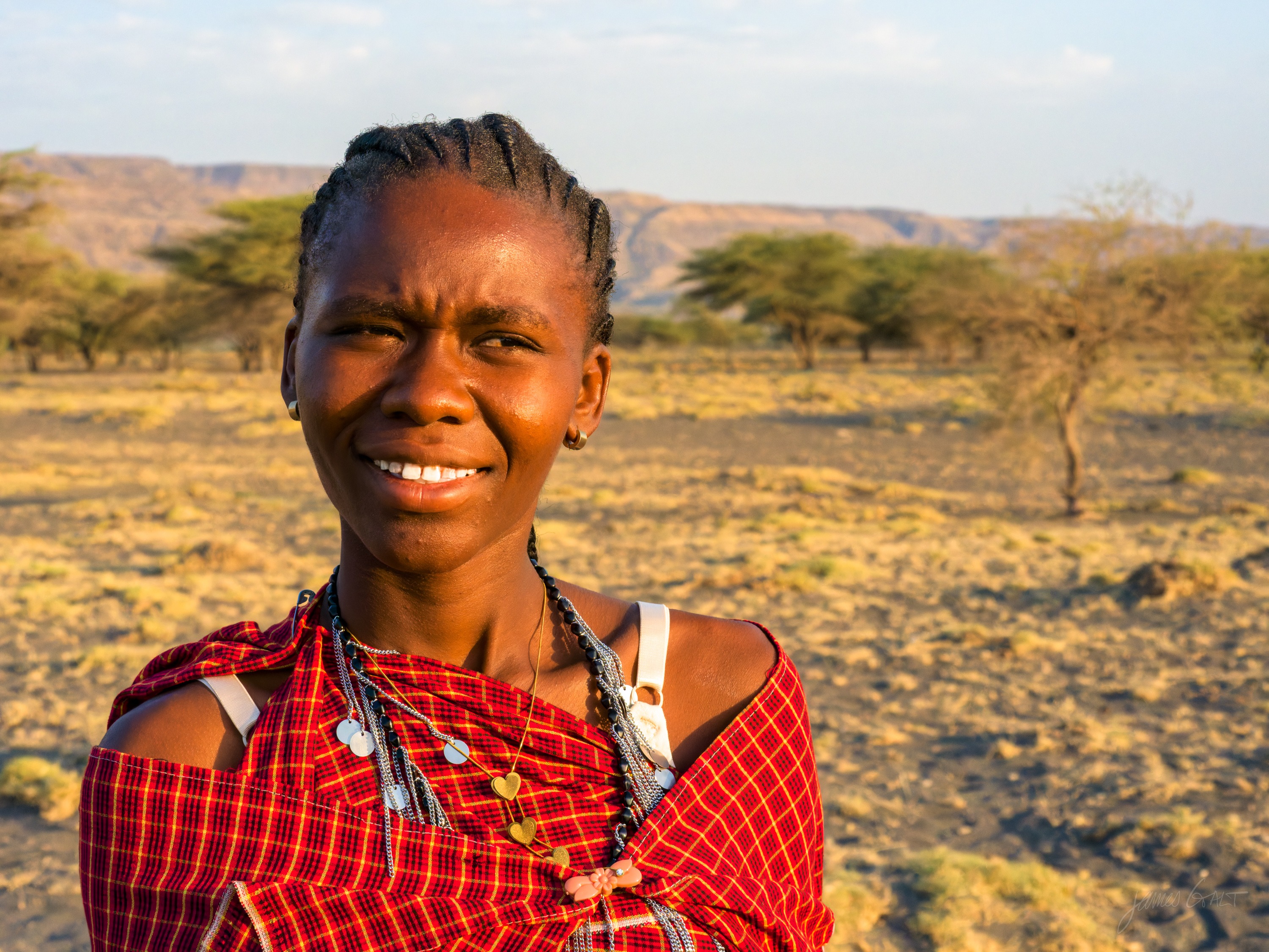 The same woman in colour, laughing, in a red beaded collar and patterned cloth, the golden grassland and small acacias behind her in low morning sun.