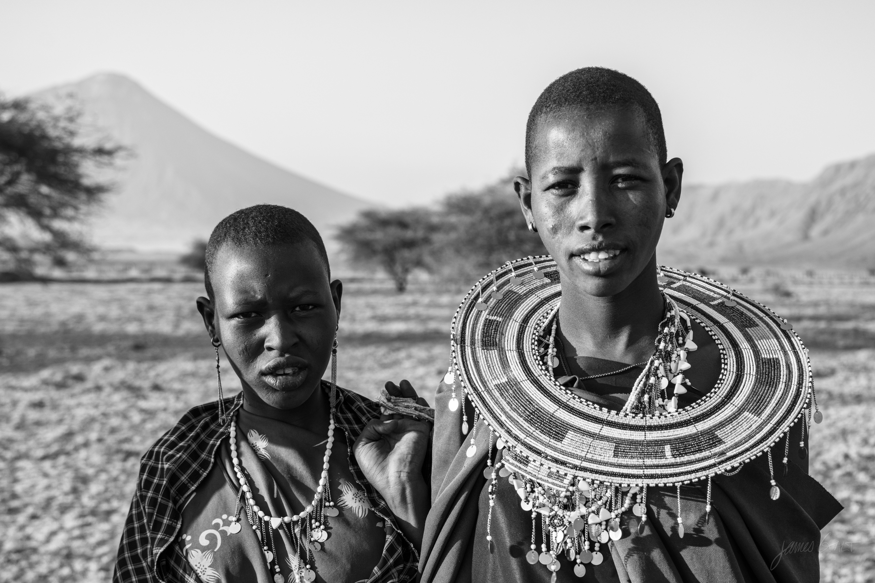 Black and white portrait of two Maasai women standing close together, both in beaded collars and headbands, a hazy volcanic cone in the far distance behind them.